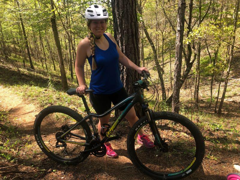 A person wearing a helmet stands next to a mountain bike on a forested trail surrounded by trees, smiling and giving a thumbs-up. They are dressed in a blue tank top and black shorts, with pink shoes, on a sunny day. Sylaward mountain bike trail.