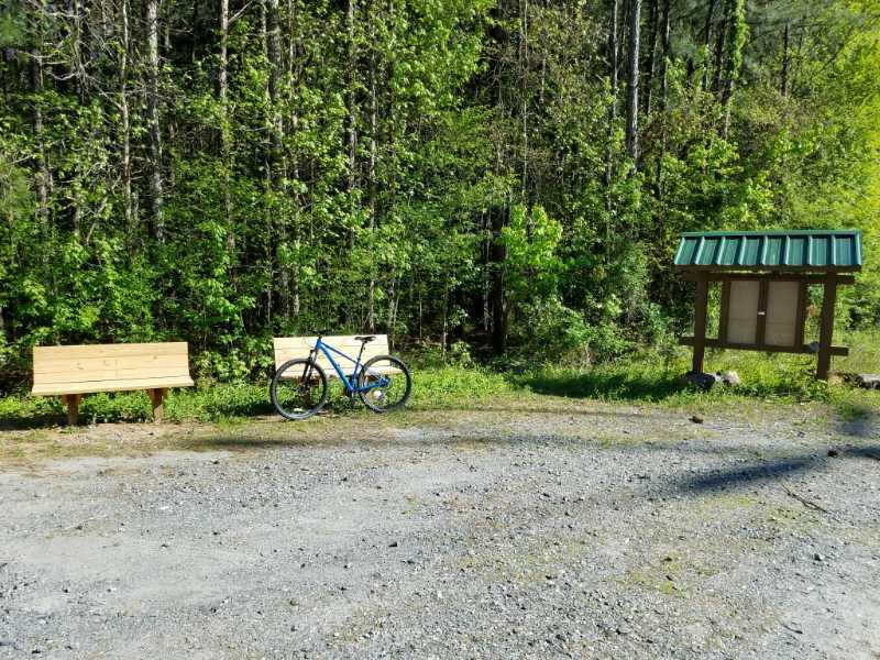 A gravel area surrounded by trees featuring two wooden benches, a blue bicycle leaning against one of the benches, and an informational signboard with a green roof. The scene is set in a lush, green forest. Austell Threadmill MTB Park (Closed) mountain bike trail.