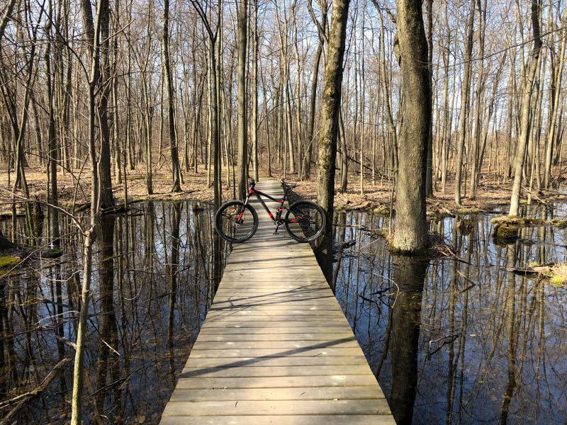 A wooden path leads through a forest, flanked by trees, with waterlogged ground visible on either side. A black bicycle is leaning against the railing of the path, reflecting the serene surroundings. The scene captures the tranquility of nature in early spring, with bare trees and clear blue skies above. Addison Oaks mountain bike trail.