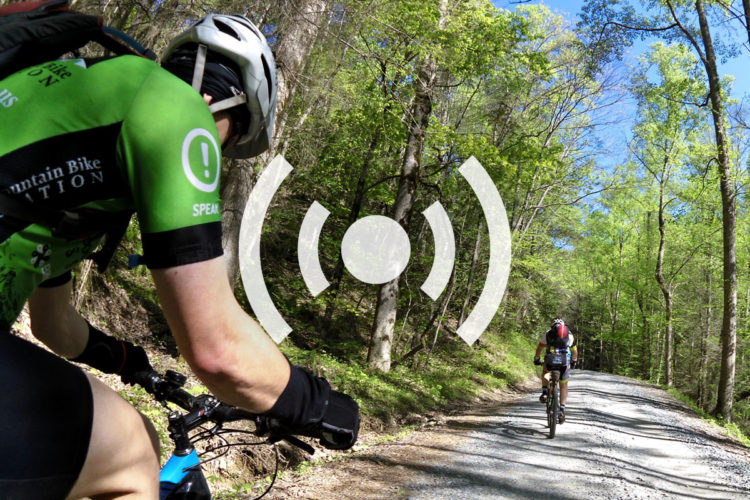 A cyclist in a green jersey rides along a dirt path surrounded by trees in a sunny, wooded area. Another cyclist is visible in the distance. An icon representing sound or communication overlays the image.