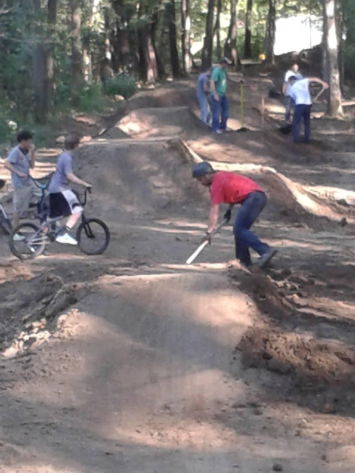A group of people working together to build a dirt bike track in a wooded area. Some individuals are digging and shaping the dirt mounds, while others observe or ride bikes nearby. The scene captures a collaborative effort to create a recreational space. Scales Lake Park Trails mountain bike trail.