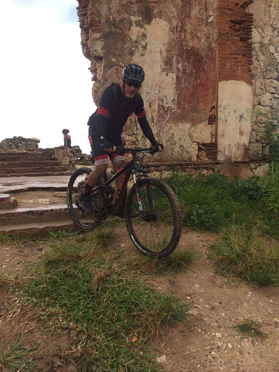 Trek Fuel EX 8 29: A cyclist in a black helmet and cycling gear rides a mountain bike on a rocky path near an ancient stone ruin, with green grass and weeds surrounding the area. In the background, a person can be seen exploring the ruins.