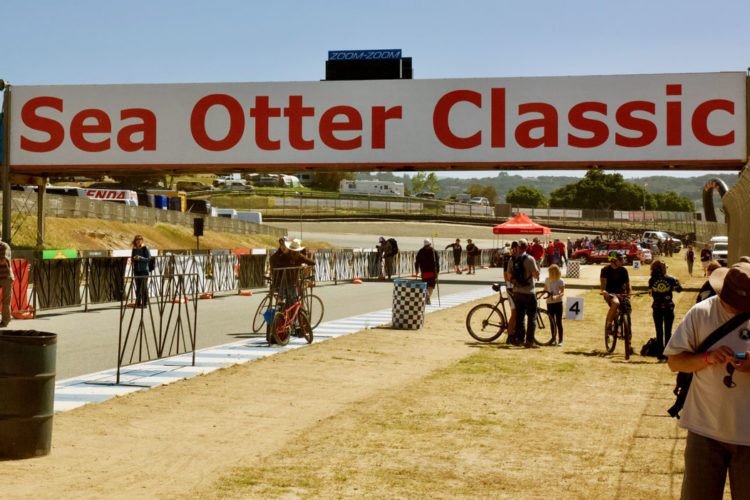 A wide shot of a cycling event venue featuring a large sign that reads "Sea Otter Classic." In the foreground, spectators and participants are gathered near the start line, with some holding bicycles. The background shows a racetrack and greenery, with tents and vehicles visible. The scene is bright and sunny, capturing the lively atmosphere of the event.