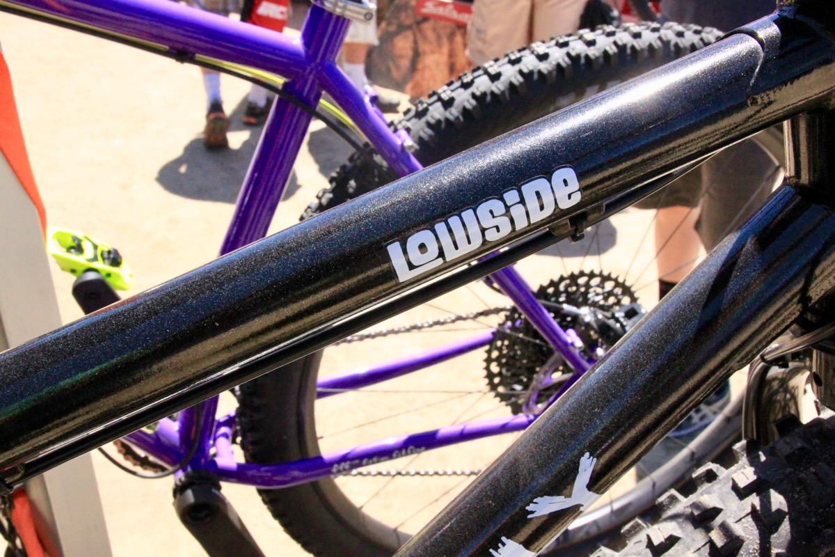 Surly Lowside: Close-up view of two mountain bike frames. The black frame prominently displays the word "Lowside" in white lettering, while a purple frame is partially visible in the background. The image captures the texture and detail of the bike frames against a blurred background of people and outdoor scenery.