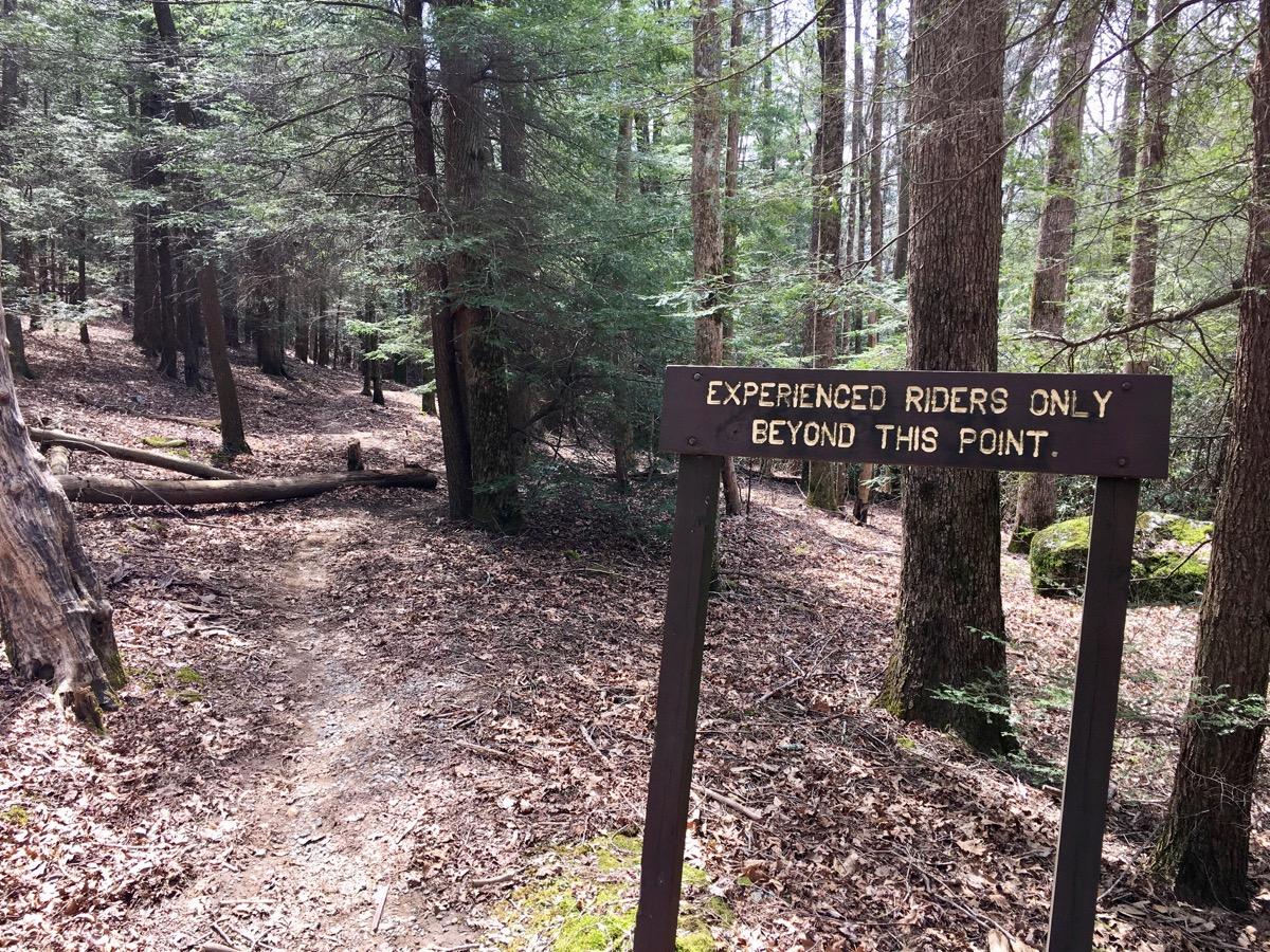 A trail sign reading "Experienced Riders Only Beyond This Point" stands in a wooded area, with a narrow dirt path leading into the trees, surrounded by fallen leaves and tall trees. Windy Gap Trail mountain bike trail.