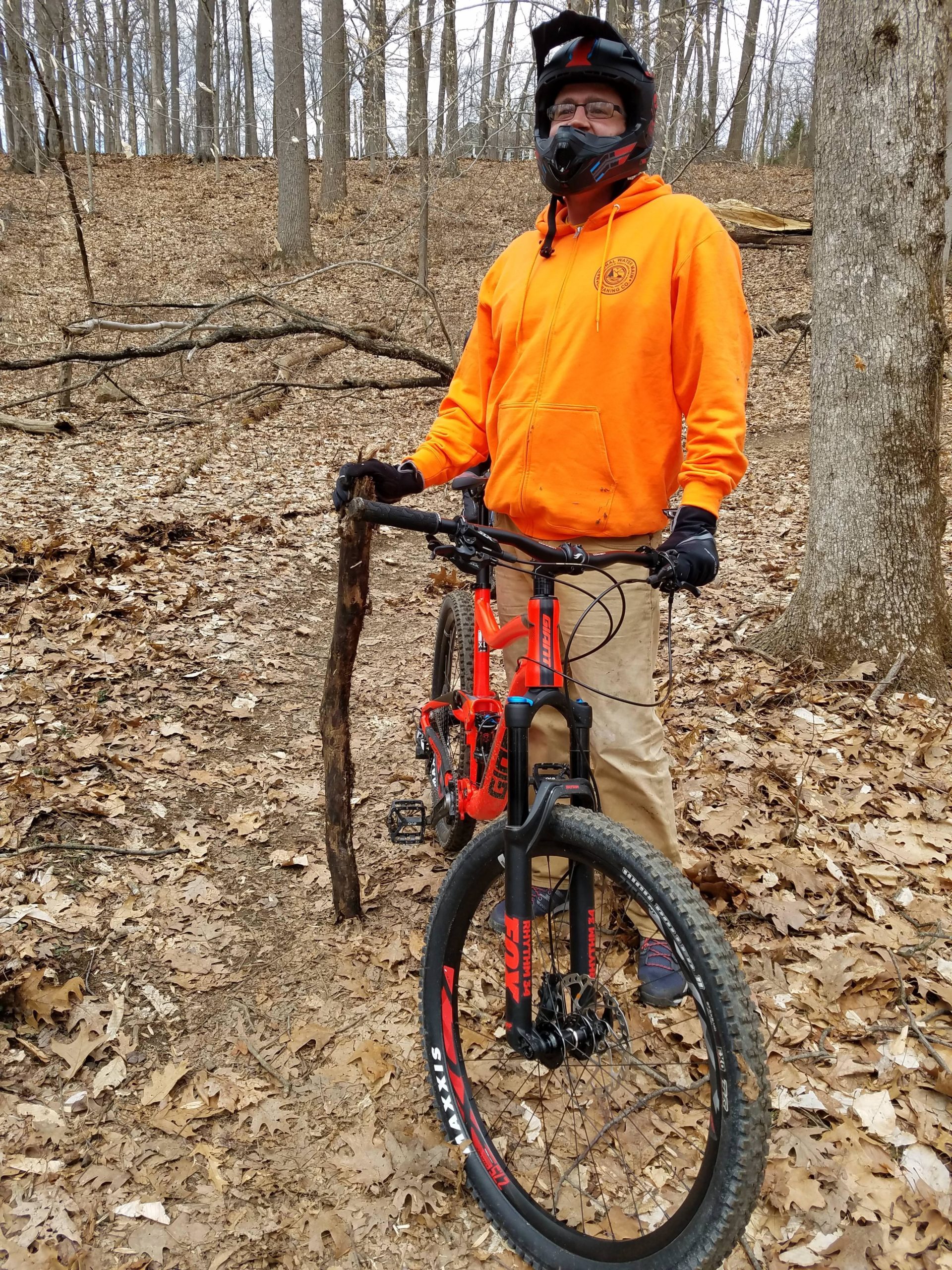 A person wearing a bright orange hoodie and black helmet stands next to a mountain bike in a wooded area. They are holding a stick and are surrounded by fallen leaves on the ground and trees in the background. The scene appears to be an outdoor biking trail. Lewis Morris mountain bike trail.