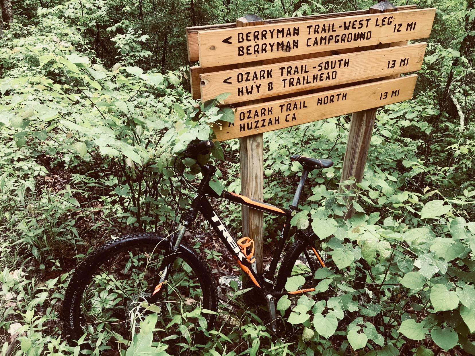 A mountain bike is leaned against a wooden trail sign in a lush green forest. The sign displays directions to various trails, including distances to Berryman Campground, Ozark Trail-South, and Ozark Trail-North. Surrounding vegetation includes dense leaves and small plants, suggesting a vibrant natural environment. Berryman mountain bike trail.