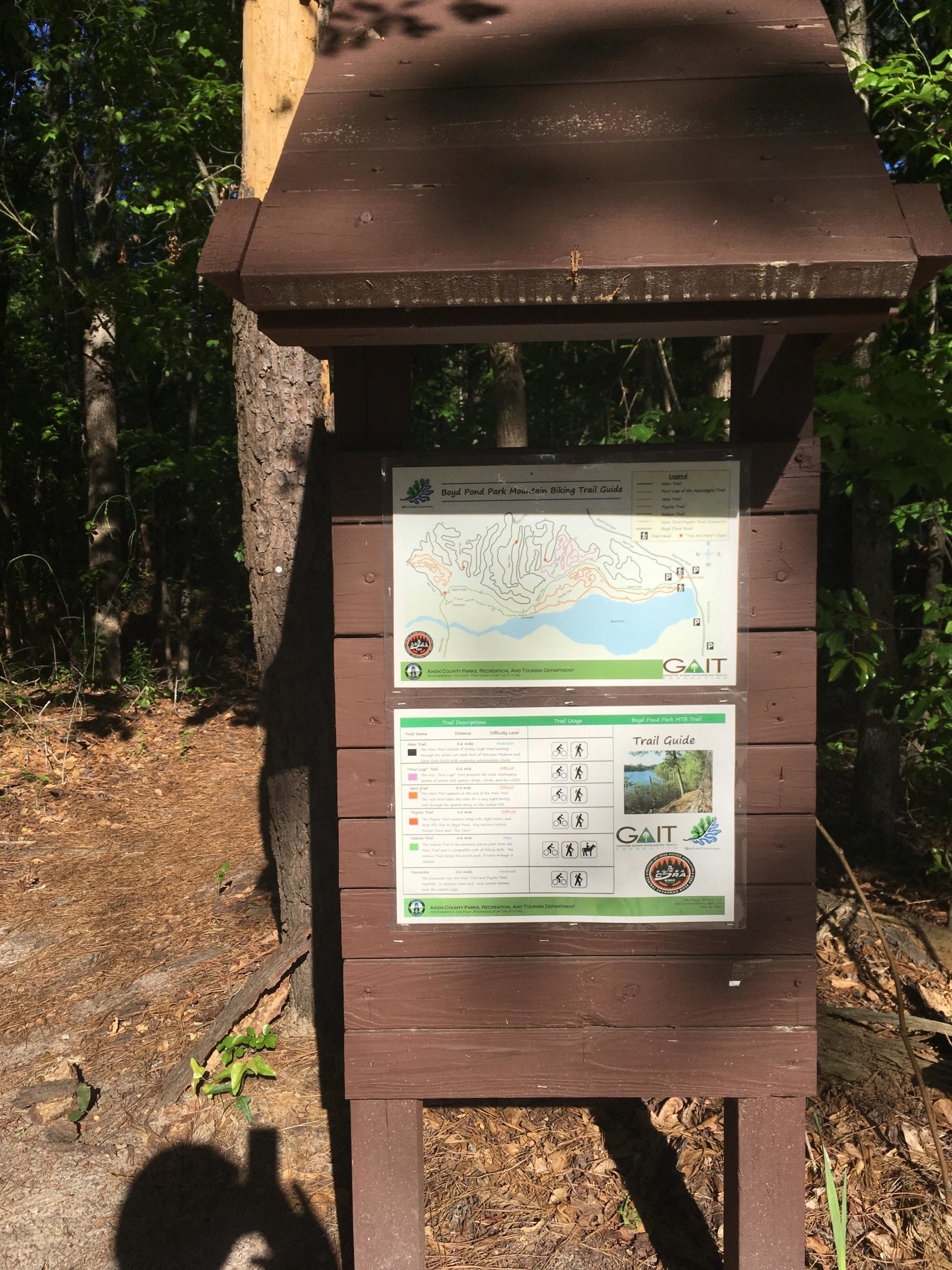 A wooden trailhead sign displaying a mountain biking trail map for Big Pond Park, surrounded by trees. The sign includes a detailed trail guide with descriptions, difficulty ratings, and icons indicating various activities, set against a backdrop of sunlight filtering through the foliage. Boyd Pond mountain bike trail.