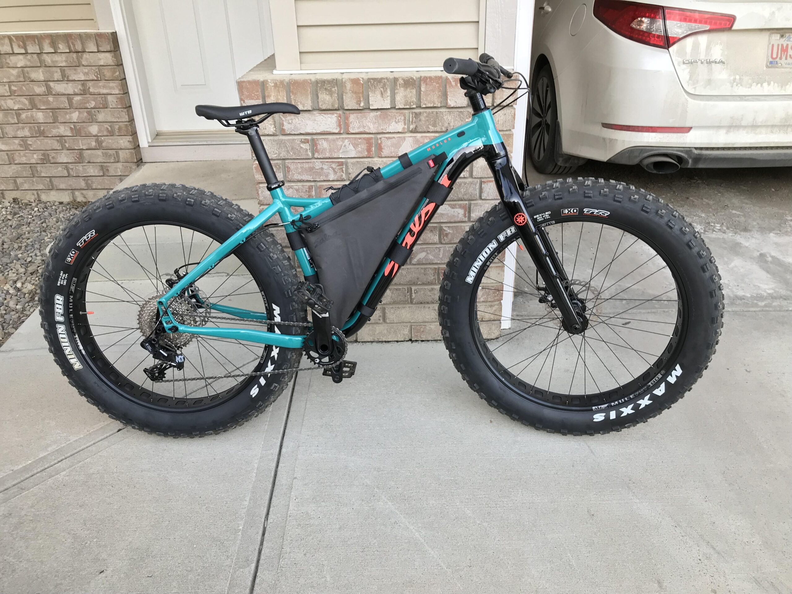 Salsa Mukluk: A teal and black fat bike is parked on a concrete surface in front of a brick wall and a door. The bike features oversized tires and a frame bag, with visible gears and pedals. In the background, a dusty car is partially visible.