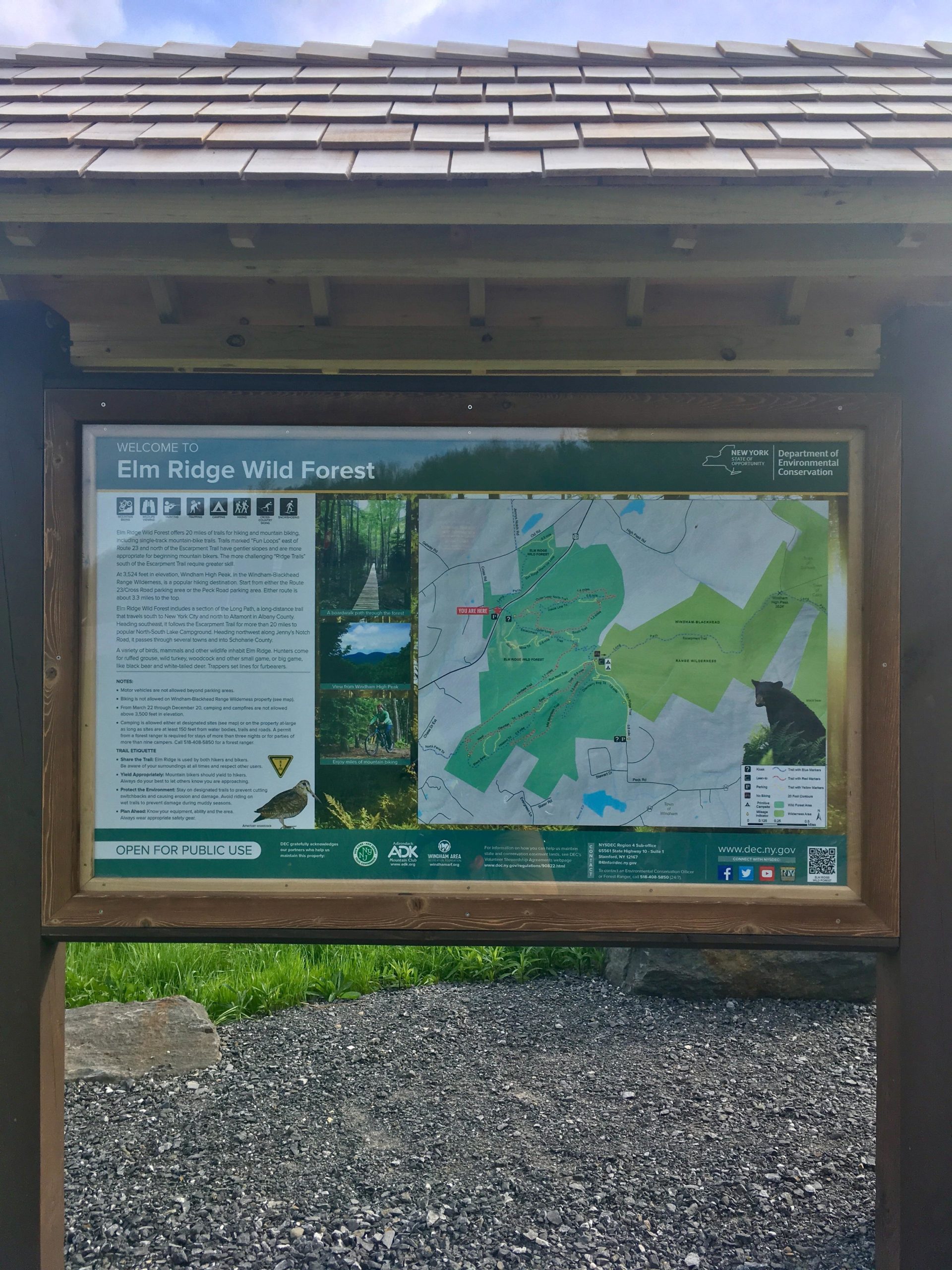 Information sign for Elm Ridge Wild Forest, featuring a map of the area with details about trails, activities, and park rules. The sign is located under a wooden shelter and includes graphics depicting outdoor activities like hiking and wildlife. The background shows green grass and a gravel path. Elm Ridge mountain bike trail.