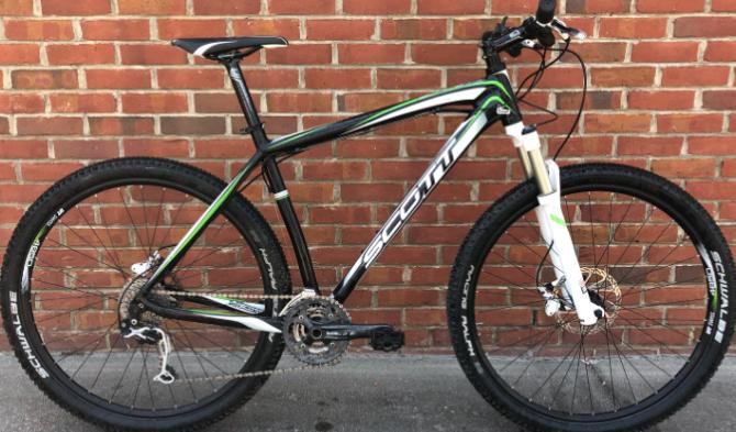 Scott Scale 29 Comp: A black and green mountain bike with a sturdy frame, front suspension, and wide tires, positioned against a brick wall. The bike features a prominent brand logo and modern components suitable for off-road riding.
