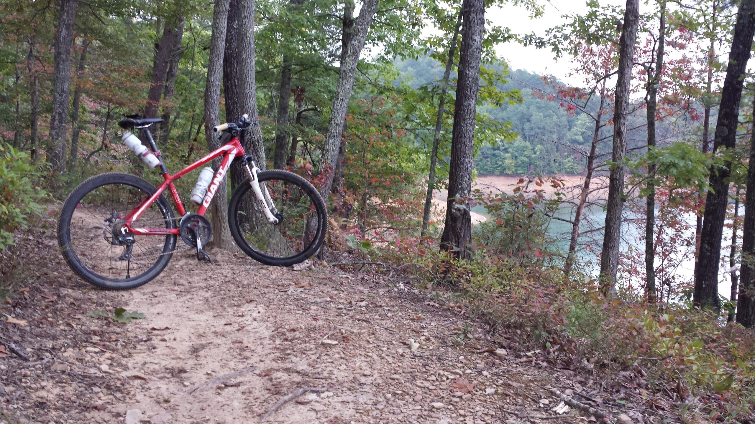 A red mountain bike parked on a dirt trail surrounded by trees, with a view of a lake in the background. The scene captures autumn foliage and a peaceful outdoor setting. Tsali Recreation Area mountain bike trail.