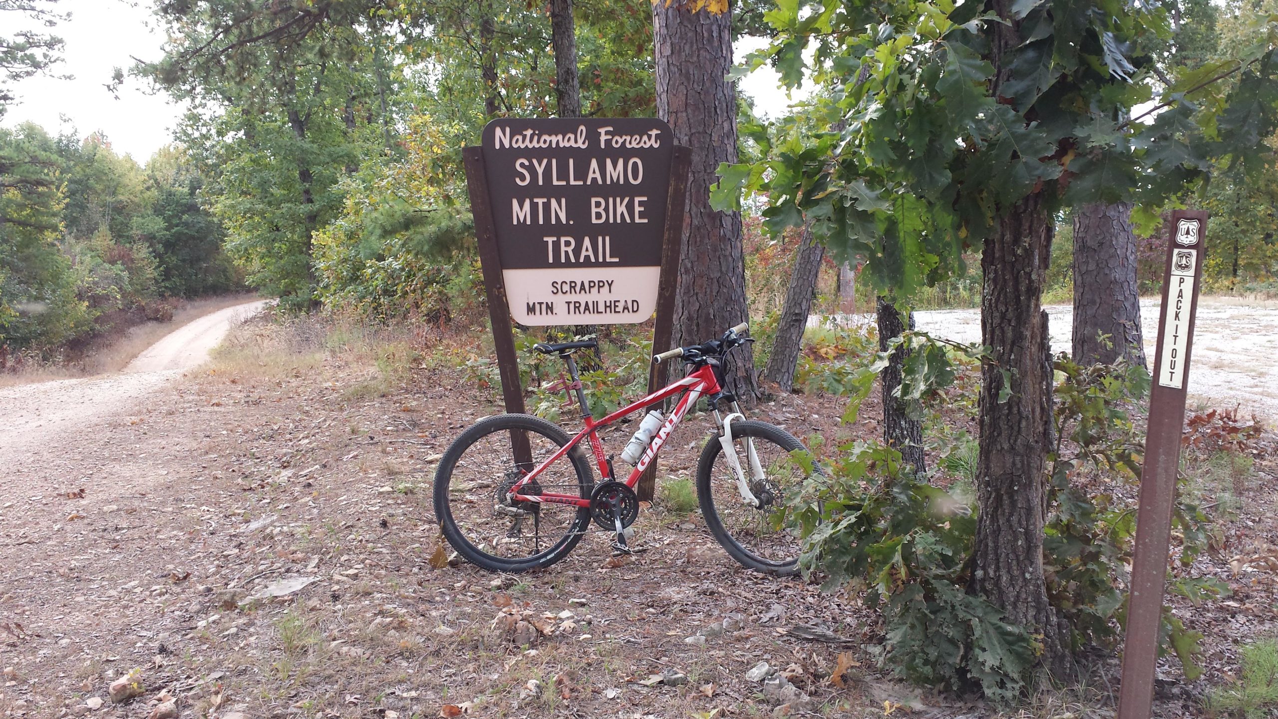 A red mountain bike rests against a sign that reads "National Forest Syllamo Mtn. Bike Trail - Scrappy Mtn. Trailhead," surrounded by trees and a dirt path in the background. There is a second sign nearby that states "Pack It Out." The scene is set in a natural outdoor environment. Syllamo Trails mountain bike trail.