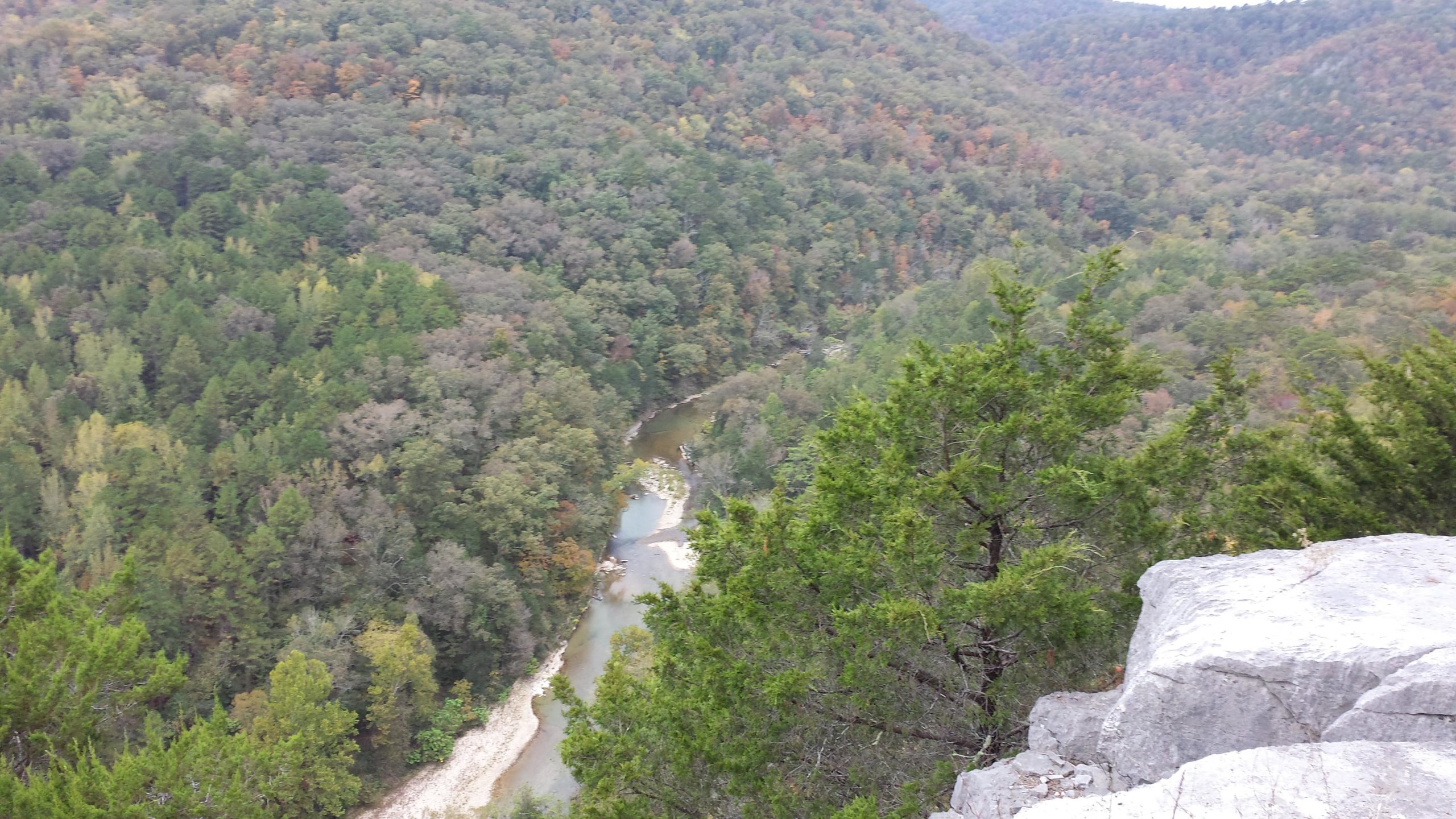 Aerial view of a lush forested landscape featuring a winding river, surrounded by trees in various shades of green and hints of autumn colors. Rocky outcrops are visible in the foreground, providing a natural vantage point over the scenery. Syllamo Trails mountain bike trail.