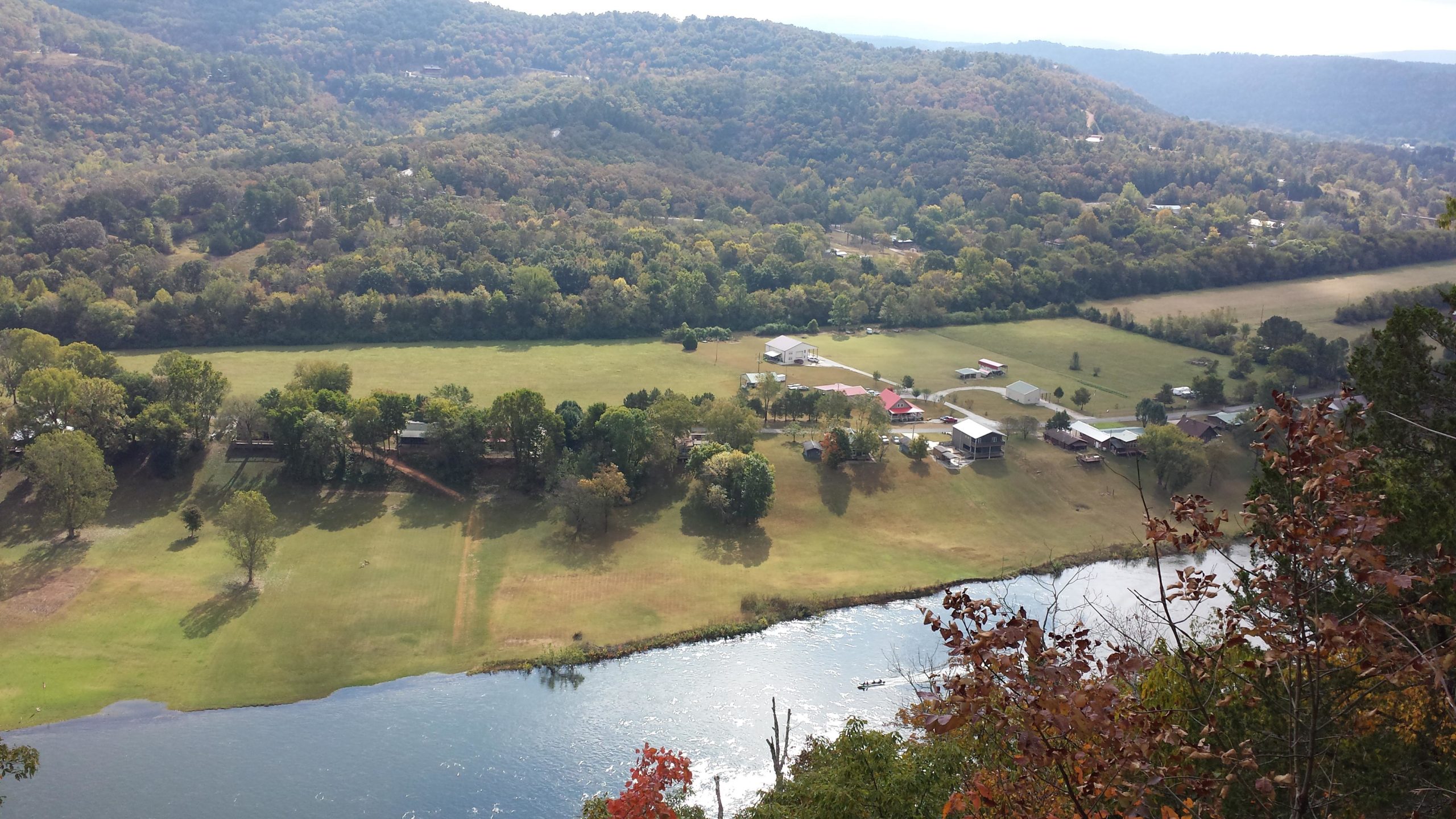 Aerial view of a winding river surrounded by lush green fields and rolling hills, with several small buildings and homes visible along the riverbank. The scene captures the natural beauty of the landscape, showcasing trees in varying shades of green, indicating a rich natural environment. Syllamo Trails mountain bike trail.