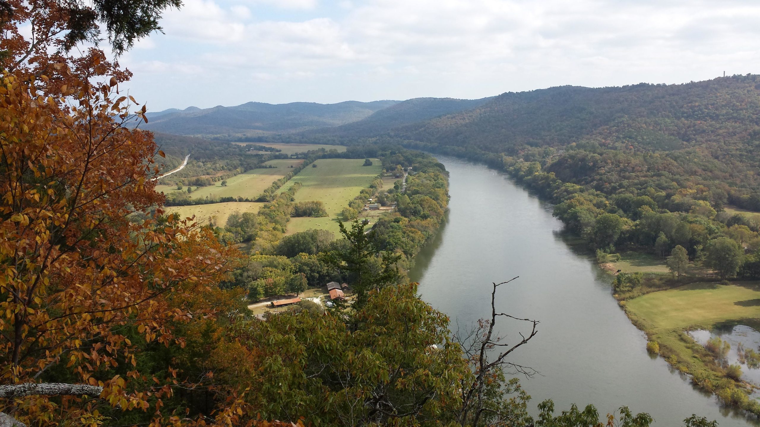 A panoramic view of a winding river surrounded by lush greenery and rolling hills, with autumn foliage in the foreground. The landscape features cultivated fields, a small cluster of buildings near the riverbank, and mountains in the background under a partly cloudy sky. Syllamo Trails mountain bike trail.
