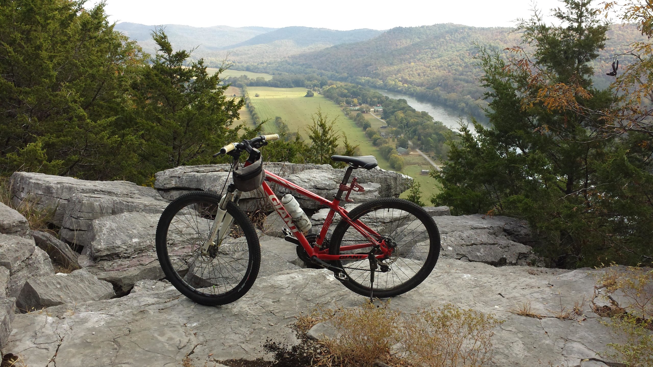A red mountain bike is resting on rocky ground, overlooking a scenic landscape of rolling hills and a winding river. The background features lush green fields and distant mountains under a cloudy sky. Syllamo Trails mountain bike trail.