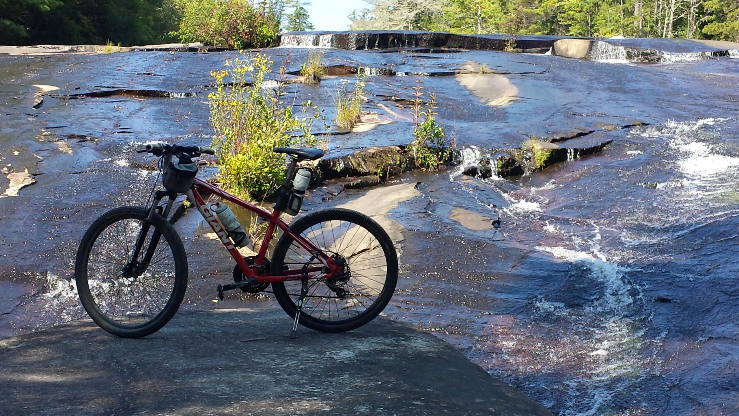 A red mountain bike positioned on a rocky surface near a flowing waterfall, surrounded by greenery and trees under clear blue skies. DuPont State Forest mountain bike trail.