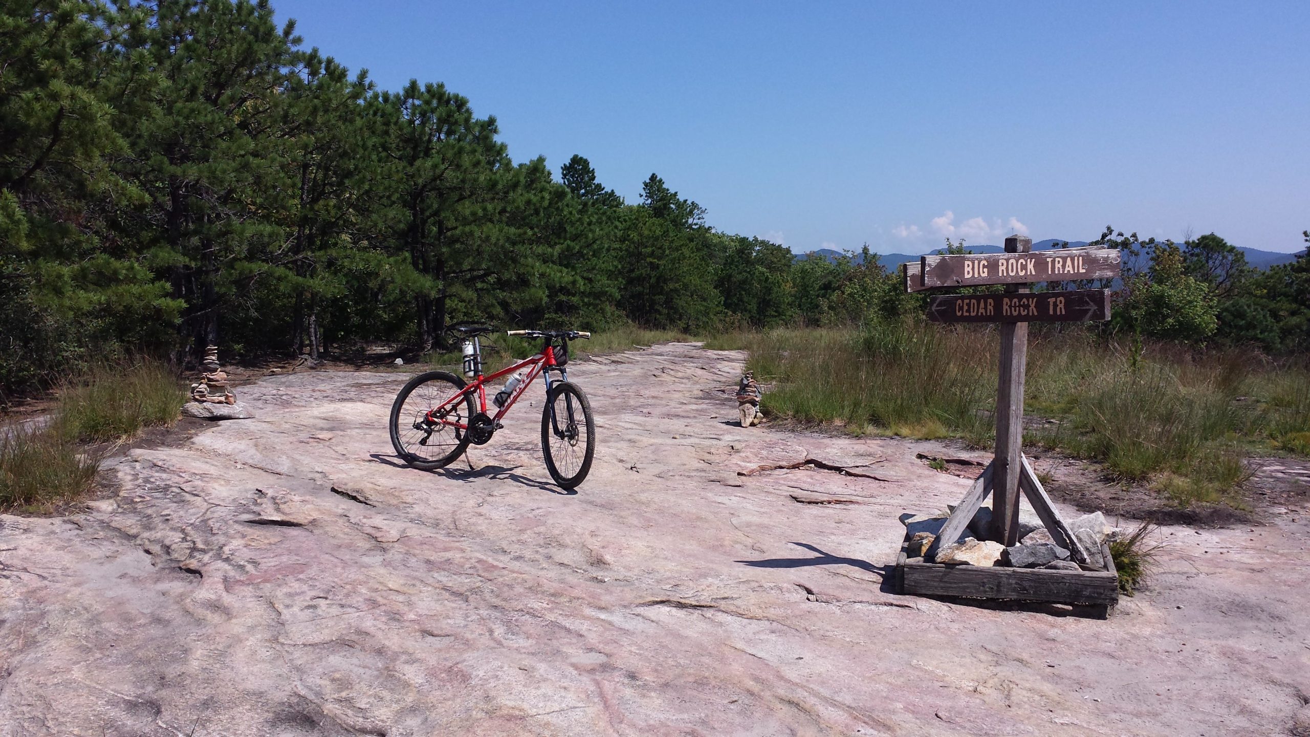 A red mountain bike is resting on a rocky trail under a clear blue sky, next to a wooden signpost indicating directions for "Big Rock Trail" and "Cedar Rock TR." The surrounding area features lush greenery and distant mountains, creating a scenic outdoor environment. DuPont State Forest mountain bike trail.