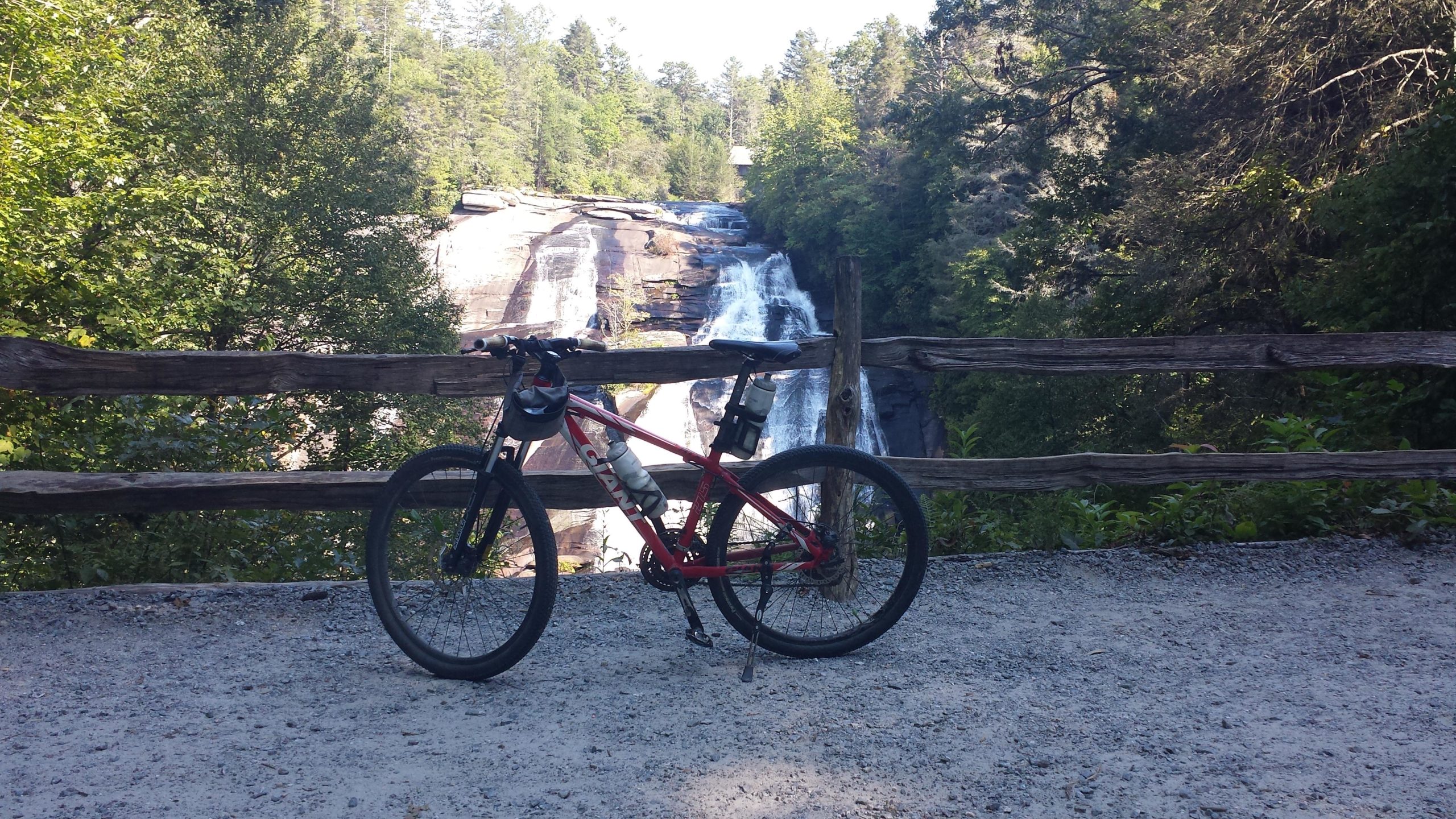 A red mountain bike parked on a gravel path near a wooden fence, with a scenic view of a waterfall and lush green trees in the background. DuPont State Forest mountain bike trail.