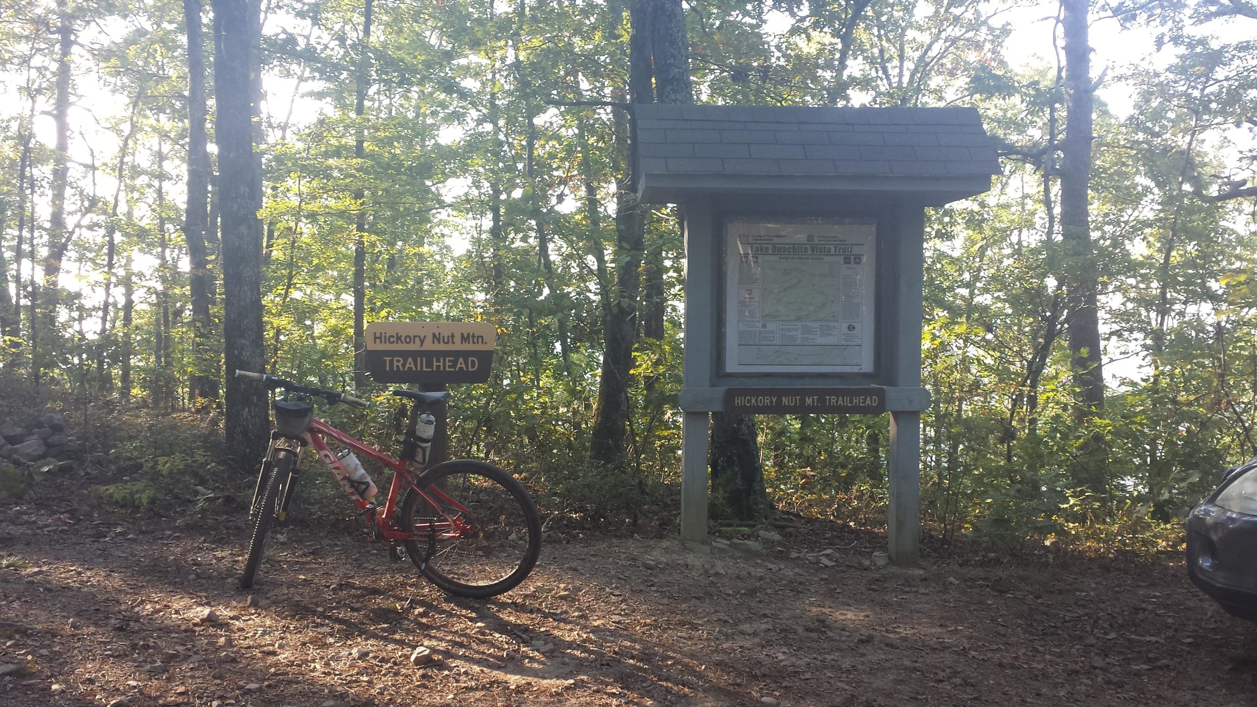 A mountain bike is parked at the Hickory Nut Mountain trailhead, which features a wooden sign indicating the trail's name. A bulletin board with a map and information about the trails is partially visible. The surrounding area is densely wooded, with sunlight filtering through the trees, creating a serene outdoor atmosphere. Lake Ouachita Vista Trail (LOViT) mountain bike trail.