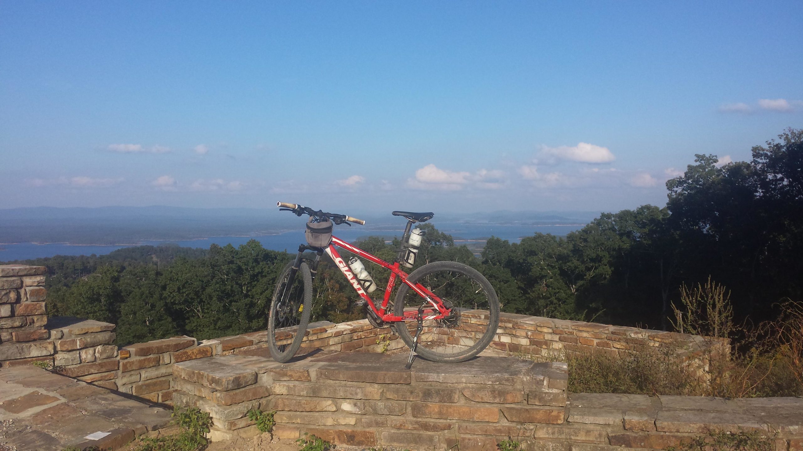 Mountain bike resting on a stone ledge with a scenic view of a lake and surrounding mountains under a clear blue sky. Lake Ouachita Vista Trail (LOViT) mountain bike trail.