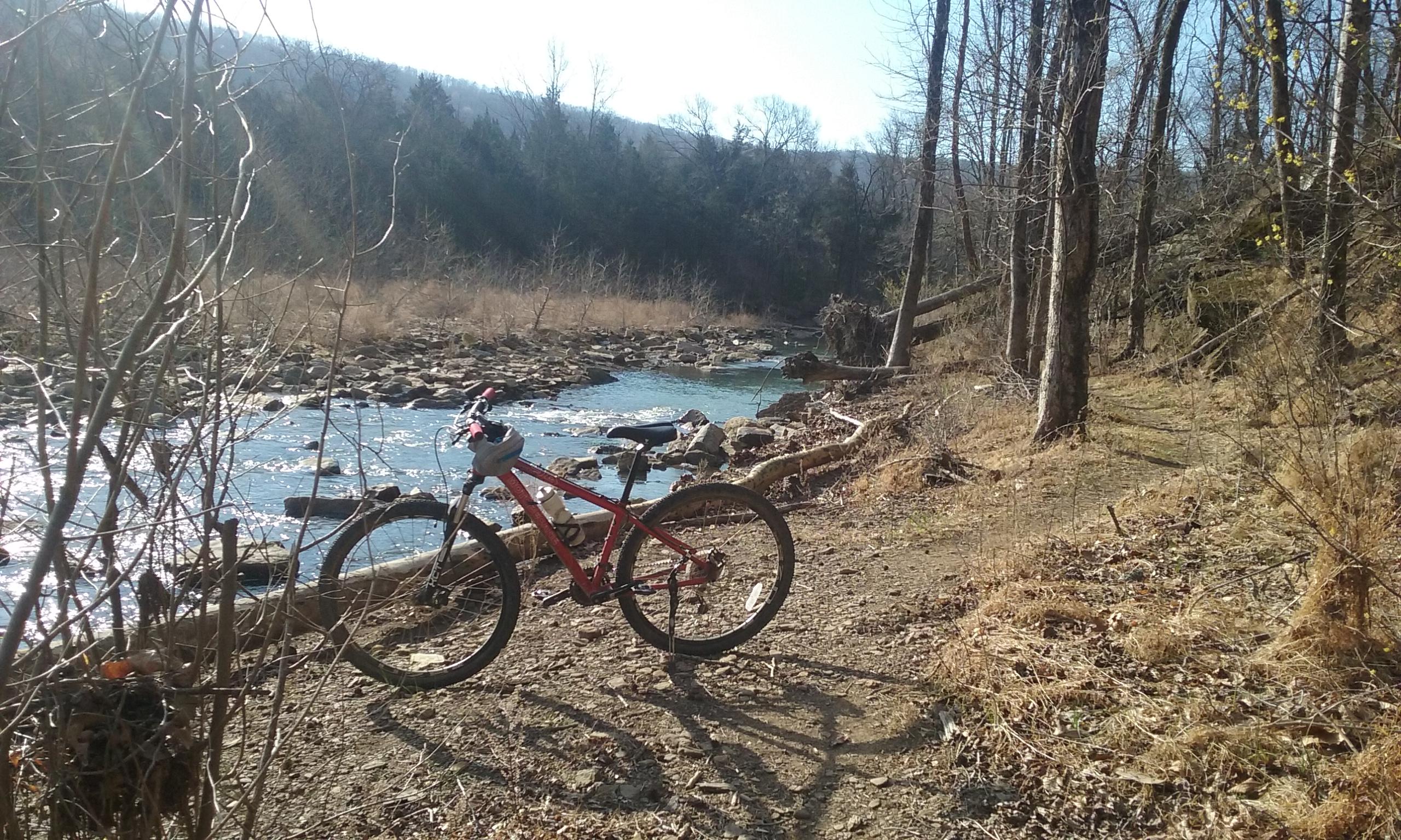 A mountain bike parked beside a flowing stream, surrounded by trees and rocky terrain. Sunlight filters through the branches, illuminating the scene on a clear day. Fossil Flats mountain bike trail.