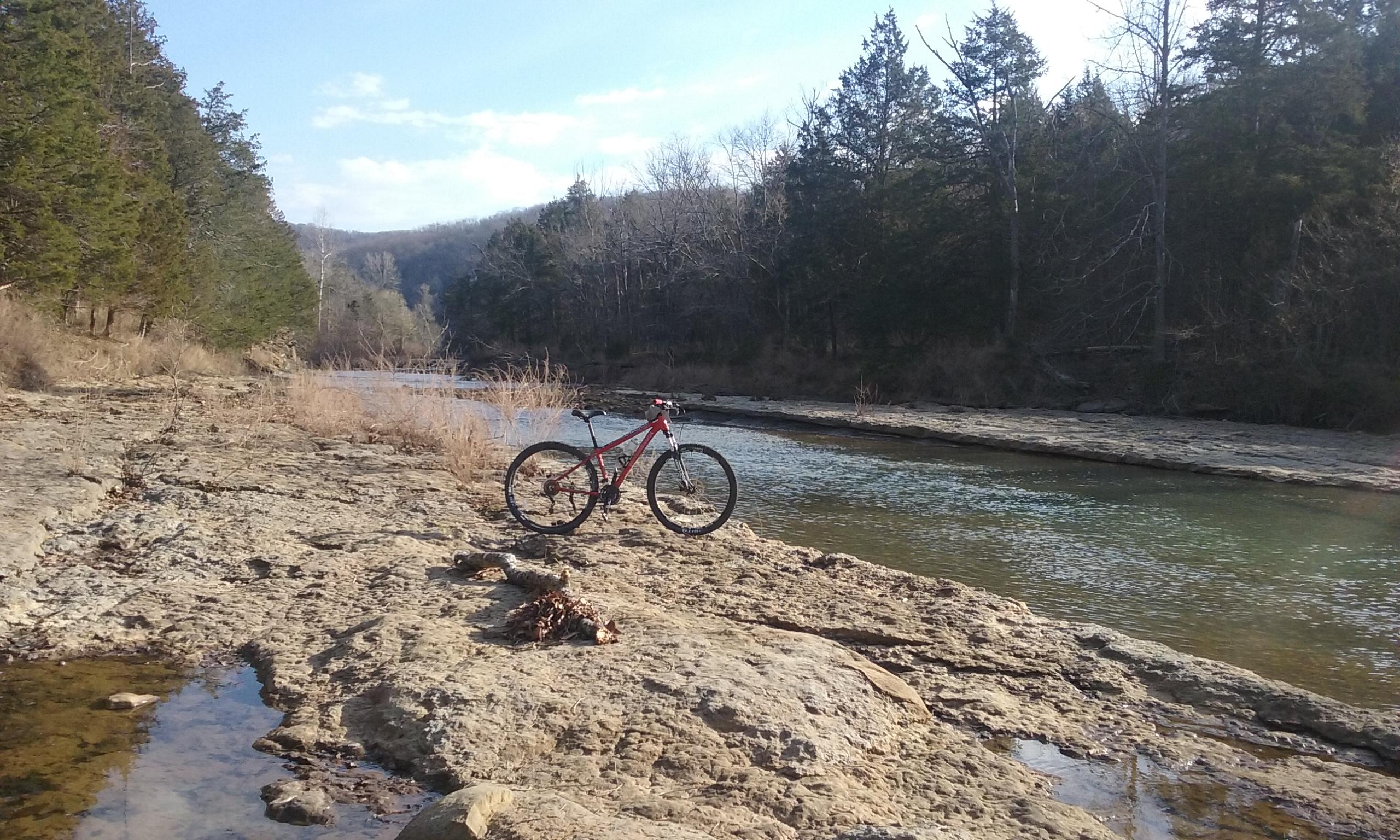 A red mountain bike rests on a rocky shoreline beside a calm river, surrounded by trees and a clear blue sky. Fossil Flats mountain bike trail.