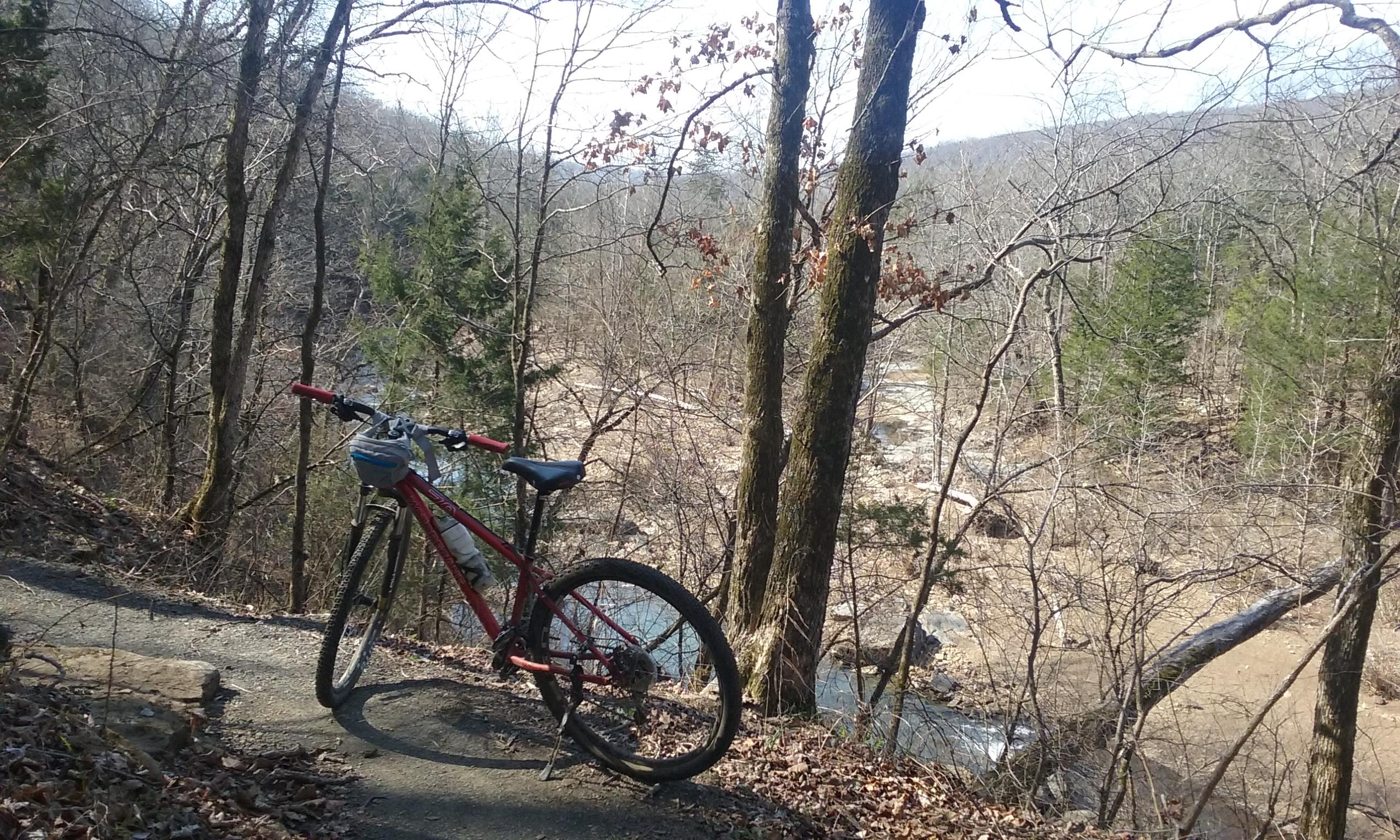 A red mountain bike rests on a gravel path surrounded by bare trees and green evergreens, with a winding river visible in the background. The scene captures a peaceful outdoor landscape during early spring. Fossil Flats mountain bike trail.