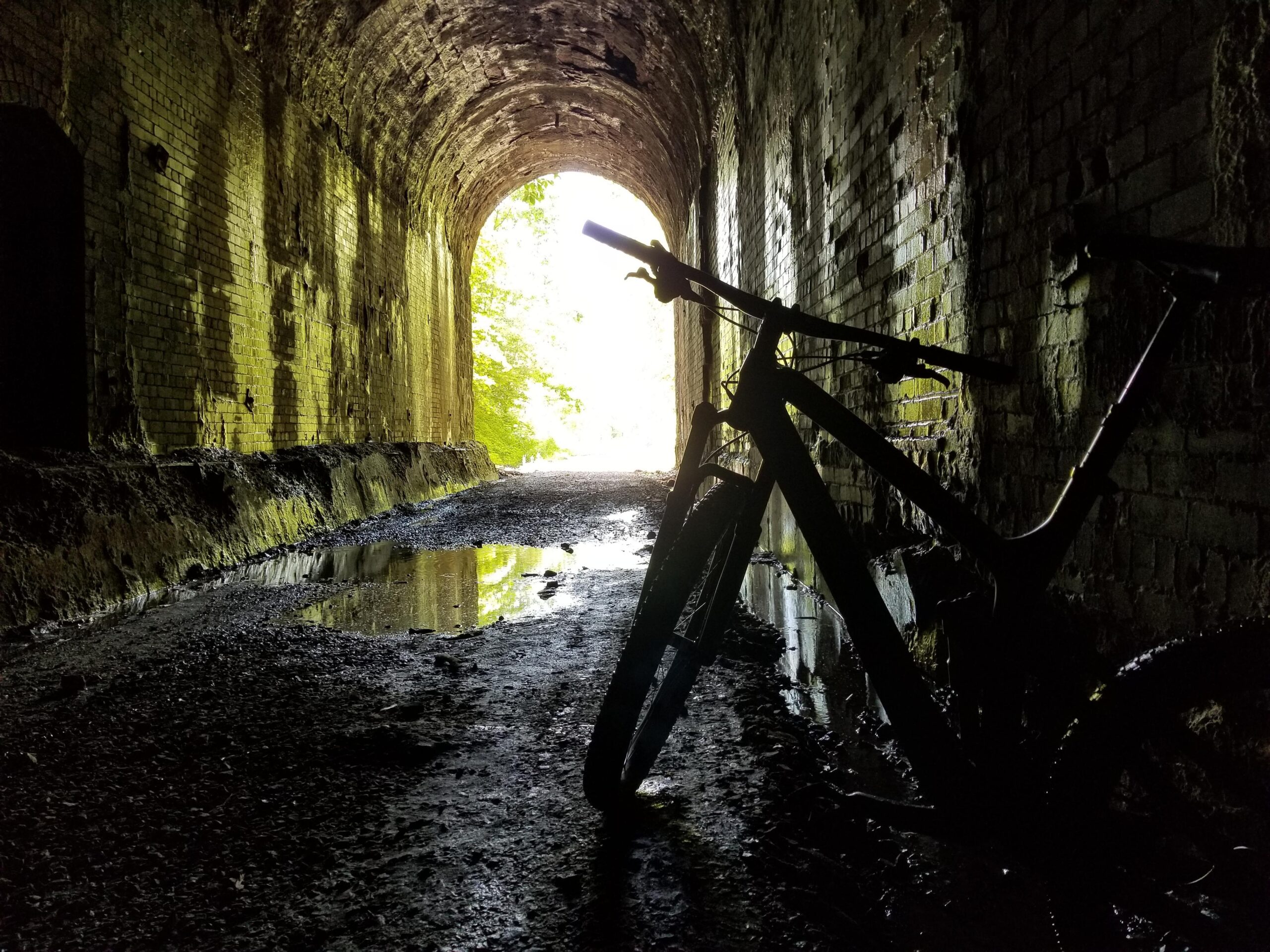 Trek Trek Fuel EX 9.8 27.5: A silhouette of a bicycle leaning against the wall of an old, moss-covered tunnel, with a bright light at the tunnel's entrance illuminating the wet ground and creating a reflective pool of water.