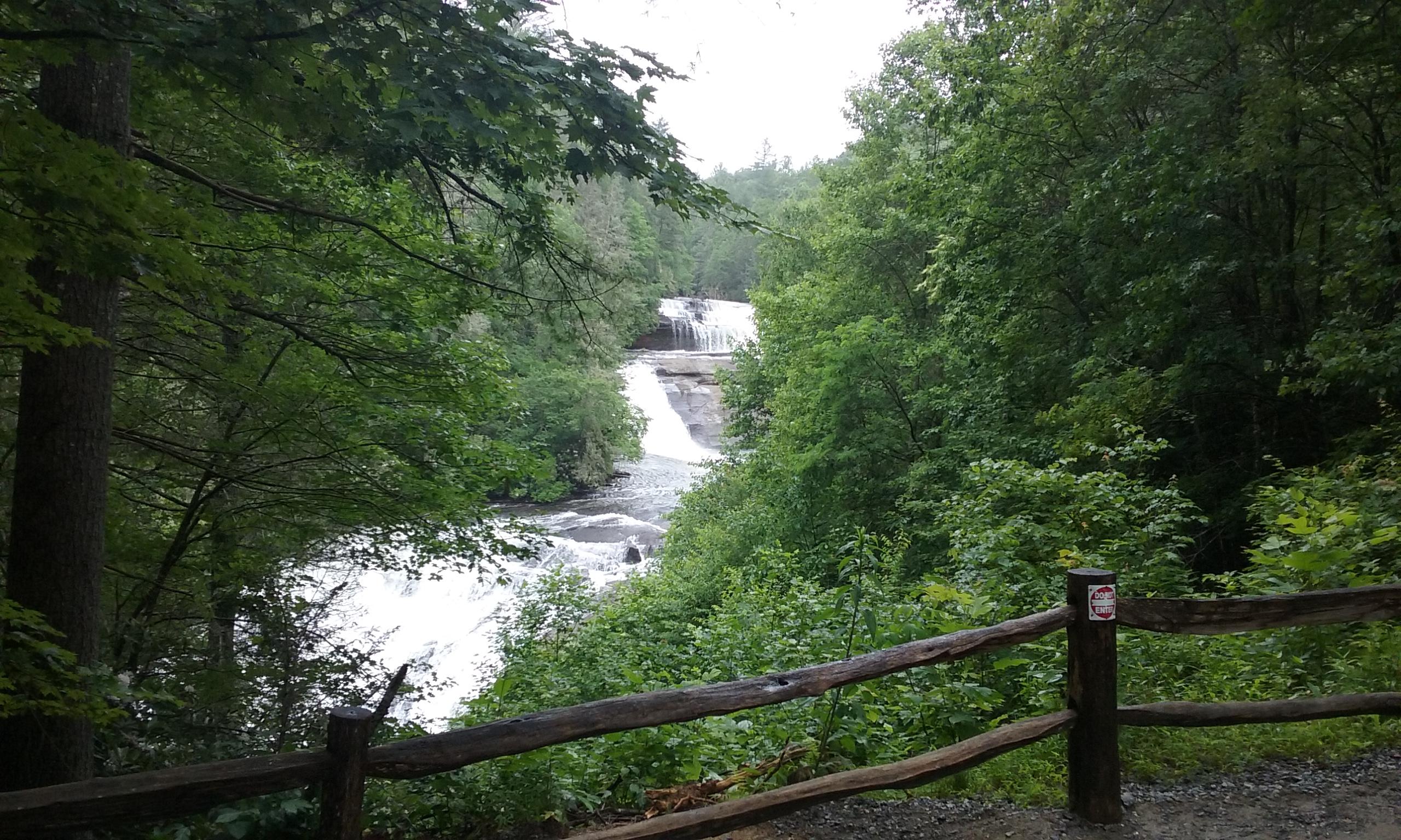 A serene view of a cascading waterfall surrounded by lush green foliage and trees, with a wooden fence in the foreground. The scene captures the beauty of nature on a cloudy day. DuPont State Forest mountain bike trail.