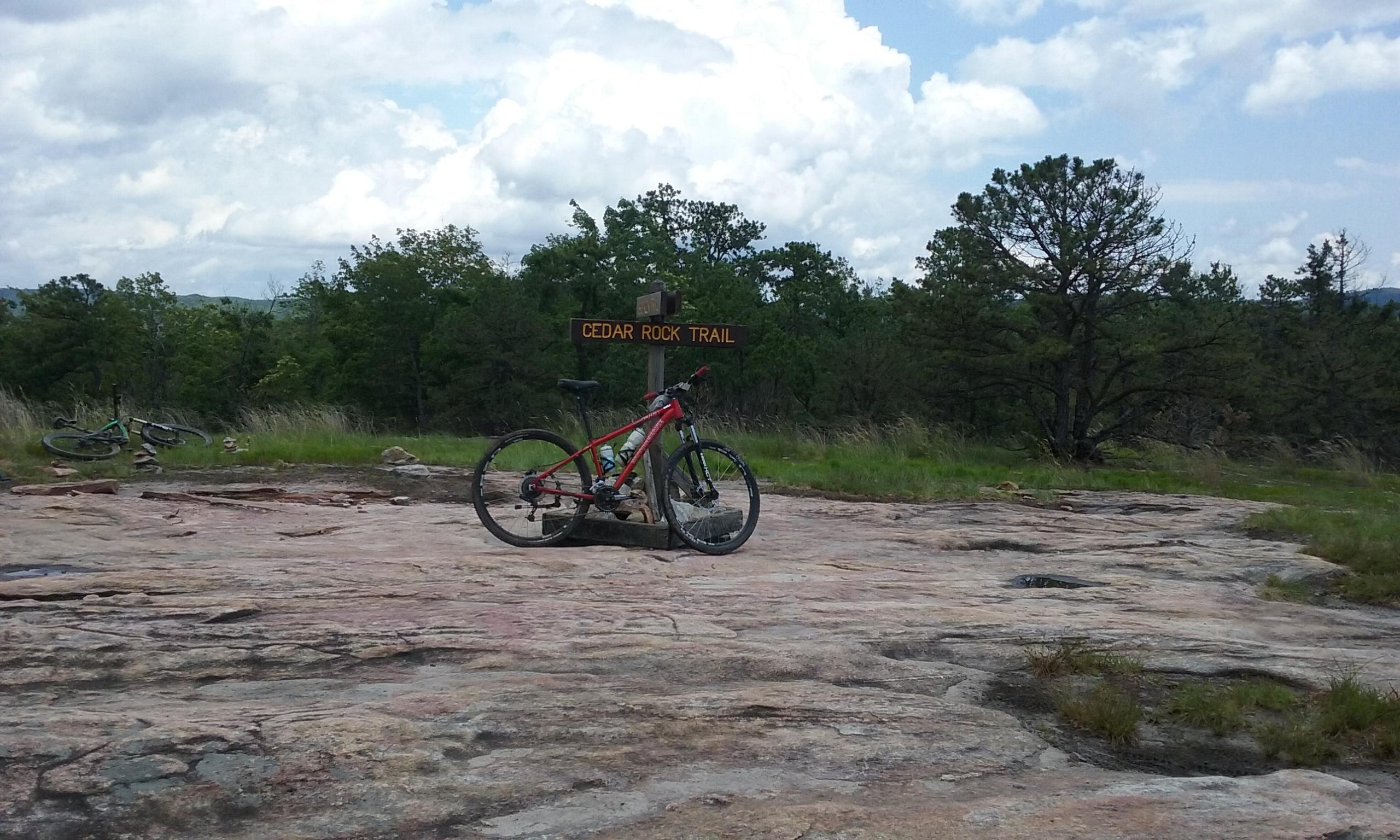 A red mountain bike rests on a rocky surface near a wooden sign that reads "CEDAR ROCK TRAIL," surrounded by green trees and a cloudy sky. In the background, another bike can be seen, blending into the natural landscape. DuPont State Forest mountain bike trail.