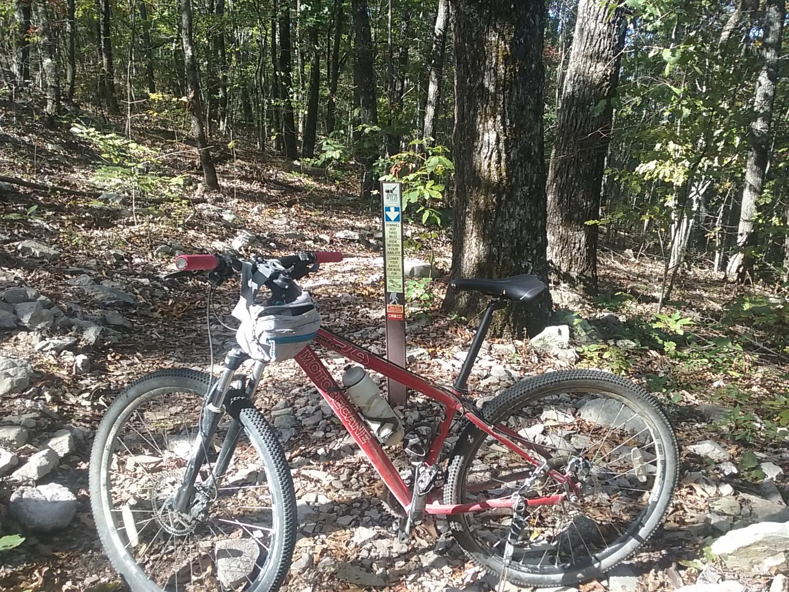 A red mountain bike with a bag attached to the handlebars is parked on a rocky trail in a wooded area. In the background, there is a trail sign partially obscured by trees, indicating the bike's location on a nature path surrounded by autumn foliage. Coldwater Mountain mountain bike trail.