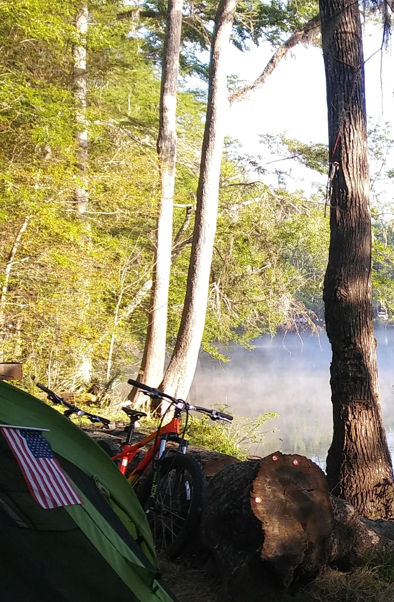 GT Ricochet: A green camping tent with an American flag emblem is set up near a serene lake, surrounded by tall trees. An orange mountain bike rests against a log, while a light mist hovers over the water, creating a peaceful outdoor scene.