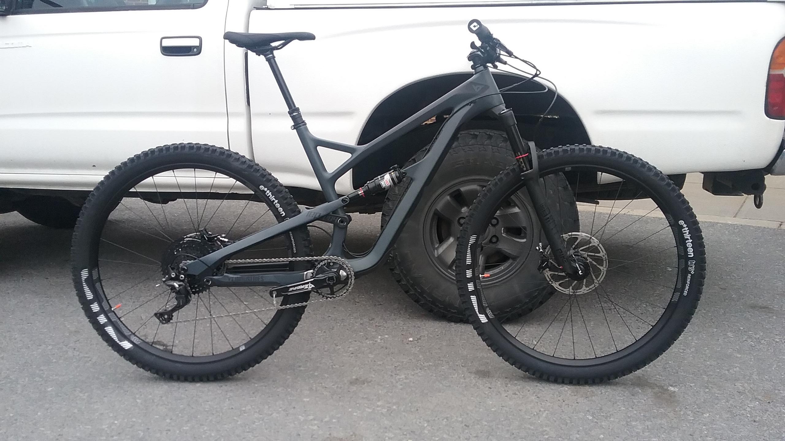 YT Industries YT Jeffsy CF: A mountain bike with a sleek gray frame and rugged tires parked next to a white pickup truck, showcasing details such as the rear derailleur and hydraulic disc brakes.