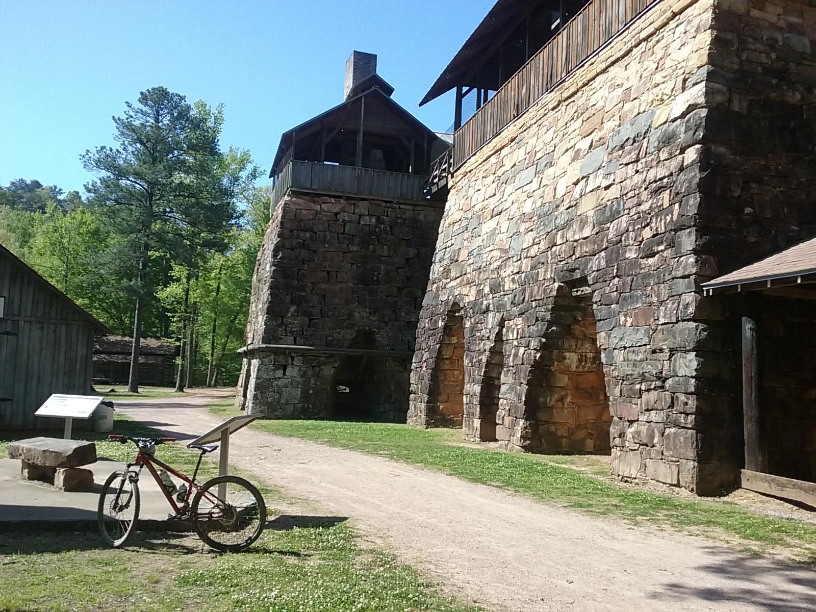 An outdoor scene featuring a stone structure with multiple arches, set against a backdrop of lush greenery and blue sky. In the foreground, a mountain bike is parked on the ground next to informational signs. The area appears to be a historical site or park, with additional smaller wooden buildings visible in the background. Tannehill Historic Ironworks State Park mountain bike trail.