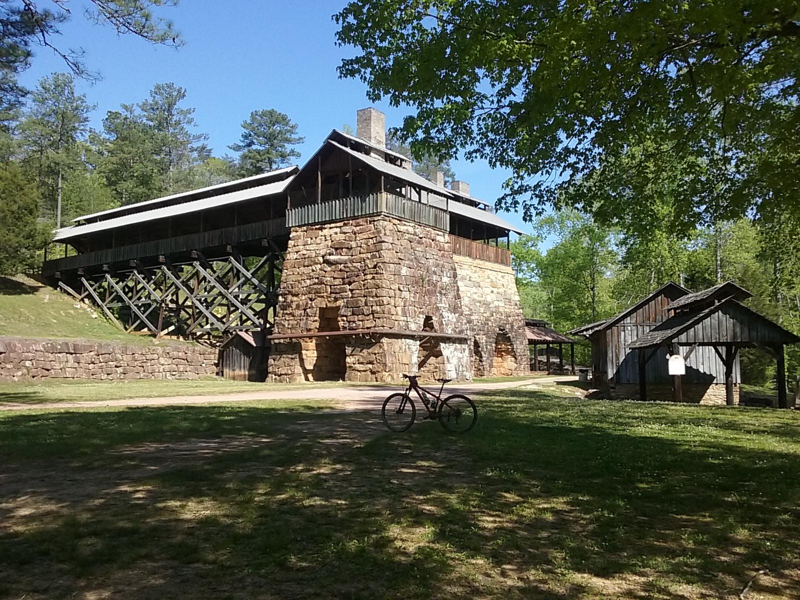 A historic stone building with wooden elements, situated amidst a green landscape. The building features a large overhanging roof and is supported by wooden beams, surrounded by trees. In the foreground, a bicycle is parked on a grassy area, with smaller wooden structures visible nearby. The sky is clear and blue, suggesting a sunny day. Tannehill Historic Ironworks State Park mountain bike trail.