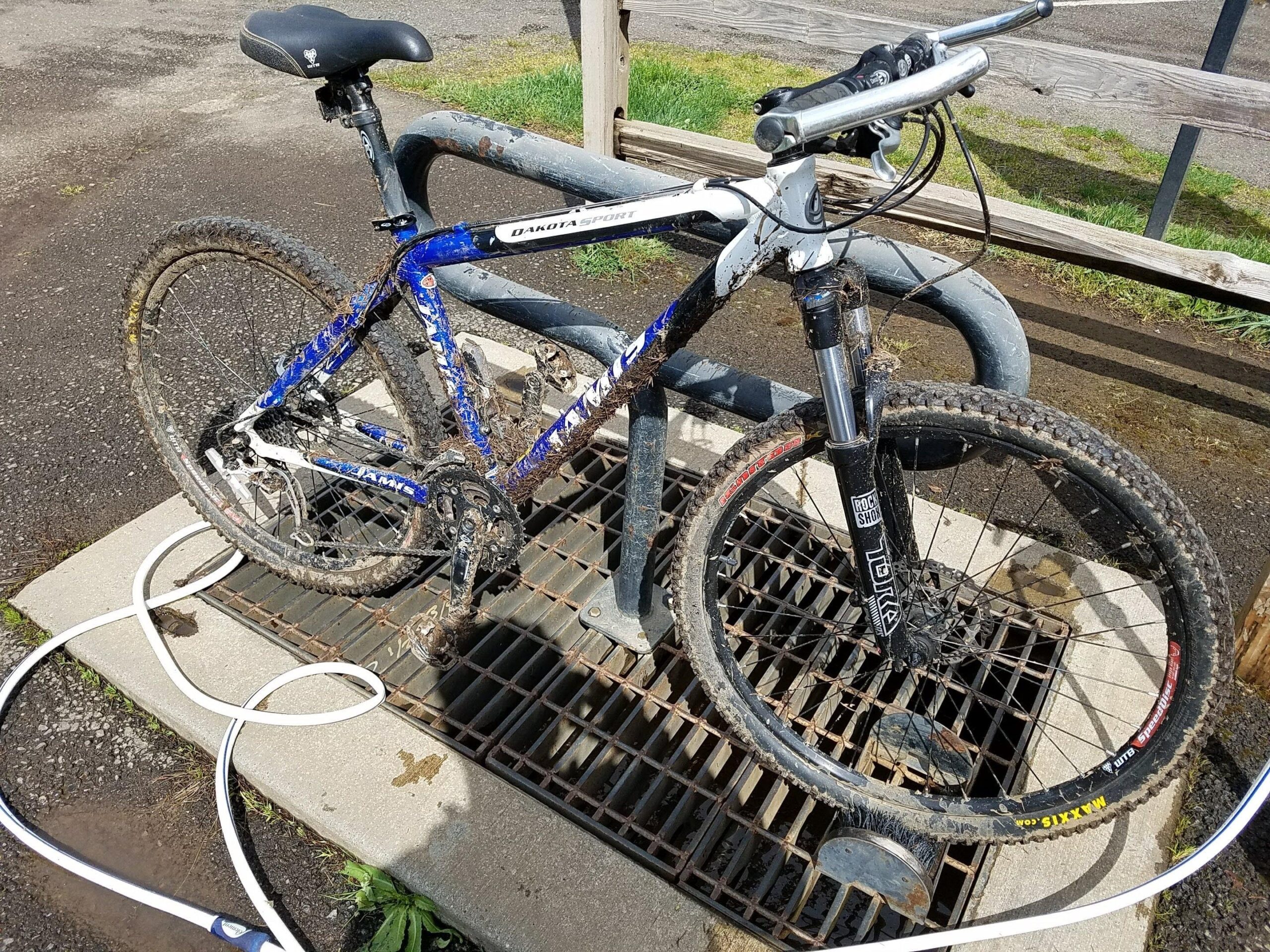 Jamis Dakota Sport: A dirty mountain bike is secured to a bike rack next to a hose on a concrete pad. The bike has muddy tires and a blue and white frame, indicating recent use on rough terrain. Fresh water puddles surround the rack on the ground.