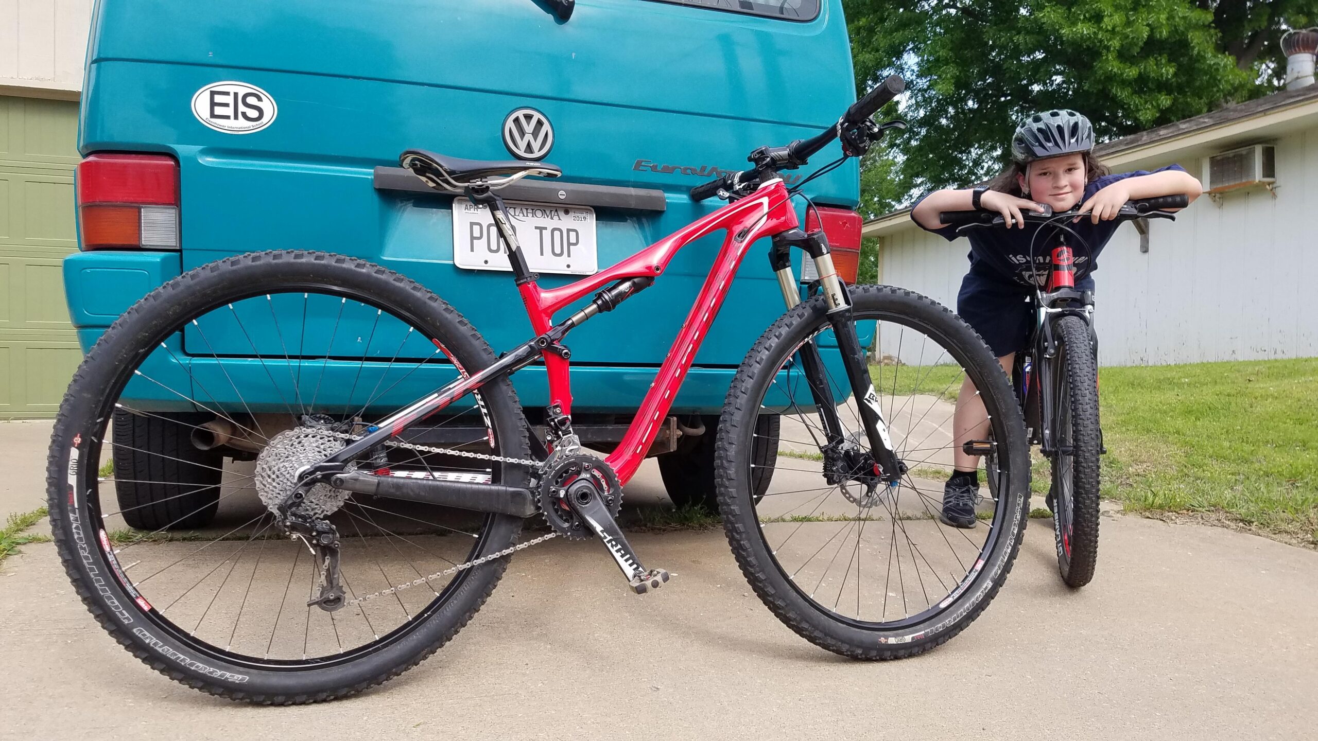 Specialized Epic Marathon 29er: A young child wearing a helmet leans on the handlebars of a black mountain bike, with a red mountain bike beside them. In the background, there is a turquoise Volkswagen van with a license plate visible. The scene is set on a driveway with green grass and a white building in the background.
