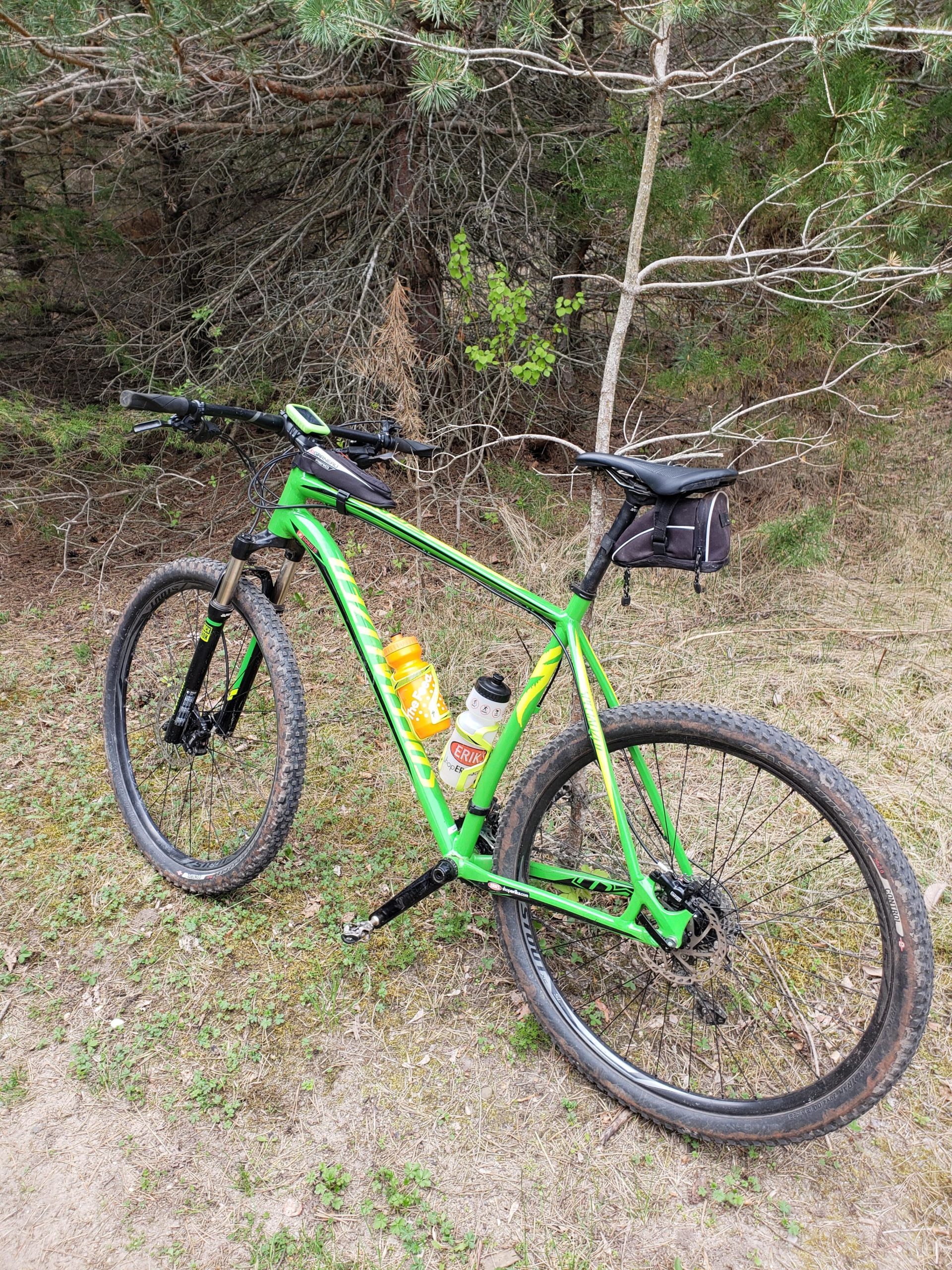 A green mountain bike parked on a dirt path surrounded by trees. The bike features a water bottle holder with two bottles attached. The terrain includes grass and small plants, indicating a natural outdoor setting. Bertram Chain of Lakes Trail mountain bike trail.