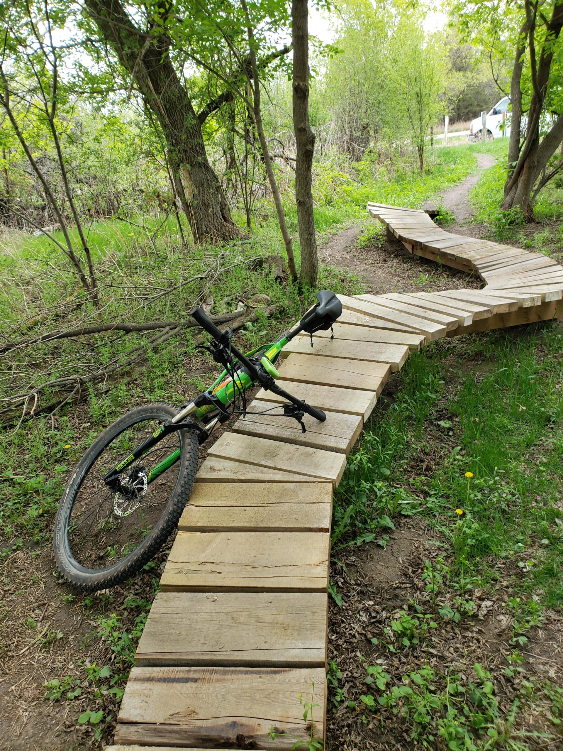 A mountain bike rests on a wooden boardwalk trail that winds through a lush, green forest. The trail is surrounded by trees and shrubs, with a hint of a pathway leading off in the distance. Dandelions and grass peek through the earthy ground, creating a vibrant natural scene. Bertram Chain of Lakes Trail mountain bike trail.