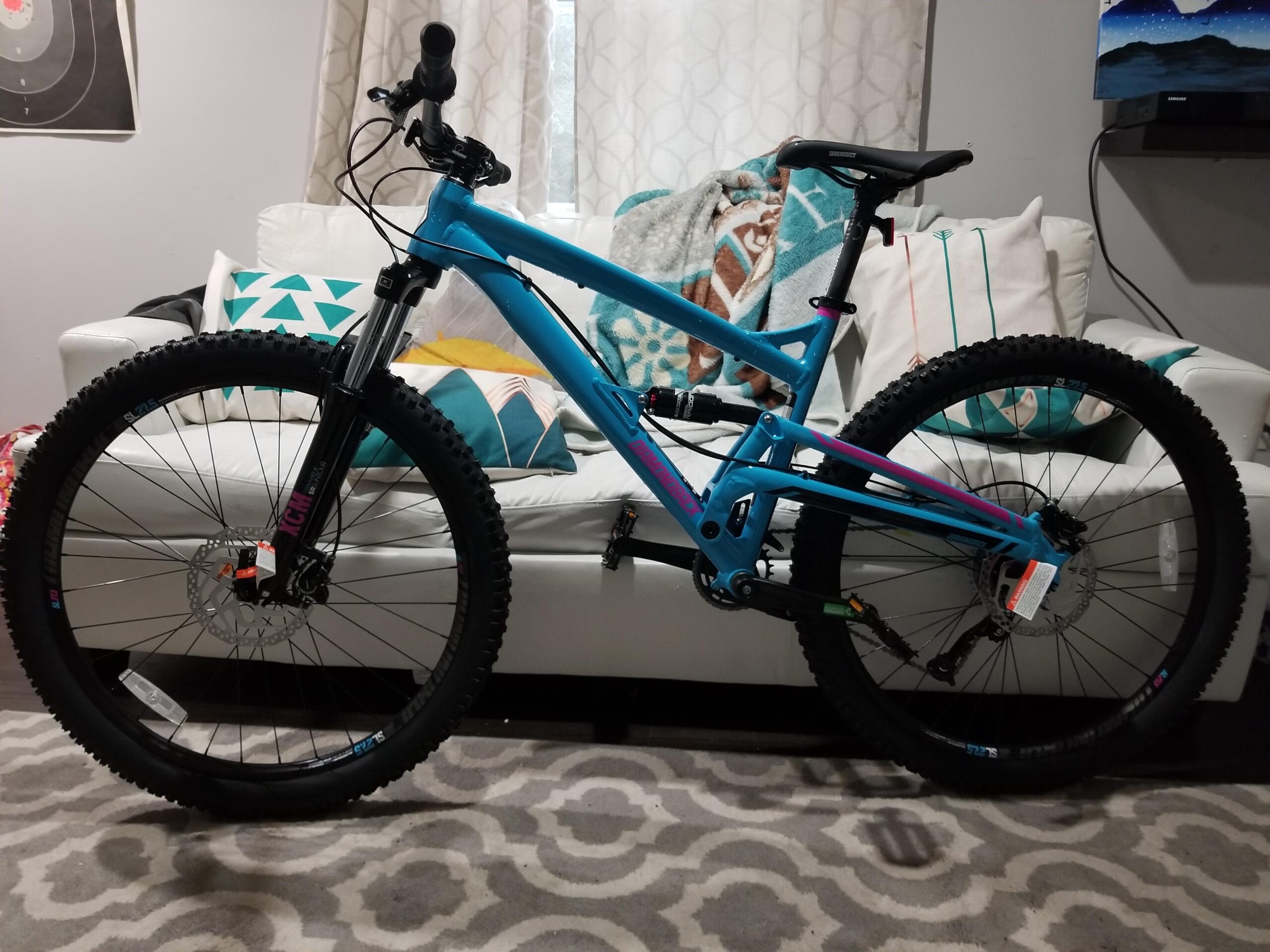 Diamondback Atroz: A bright blue mountain bike is positioned in a living room setting, leaning against a white sofa adorned with colorful cushions. The bike features thick tires and a sturdy frame, showcasing its design for off-road riding. Soft light filters through sheer curtains, illuminating the cozy space with a patterned rug on the floor.