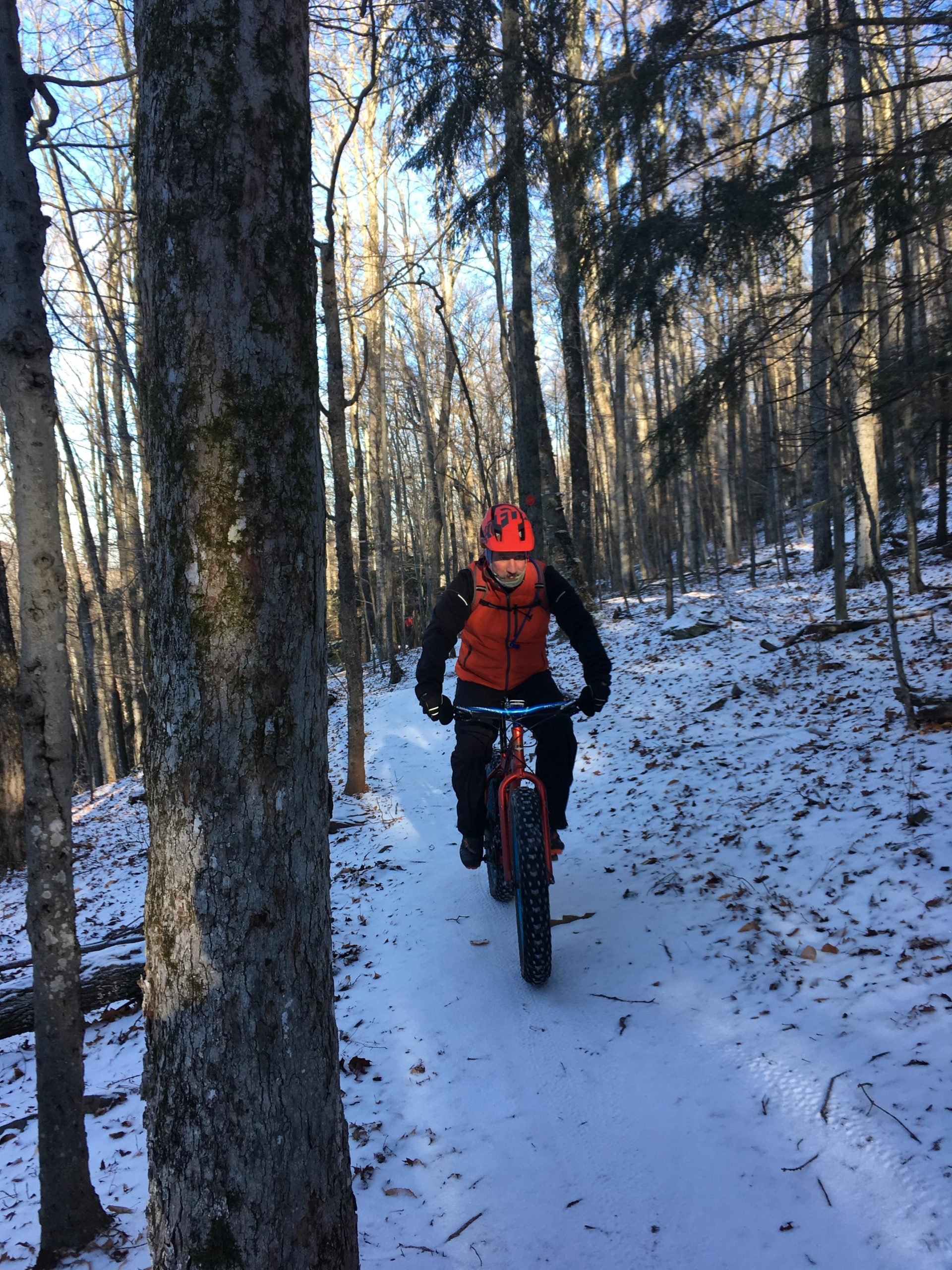 A person riding a fat tire bike on a snowy trail surrounded by trees in a winter forest. The rider is wearing an orange jacket, black gloves, and a helmet, while the path is lined with snow and bare branches. Elm Ridge mountain bike trail.