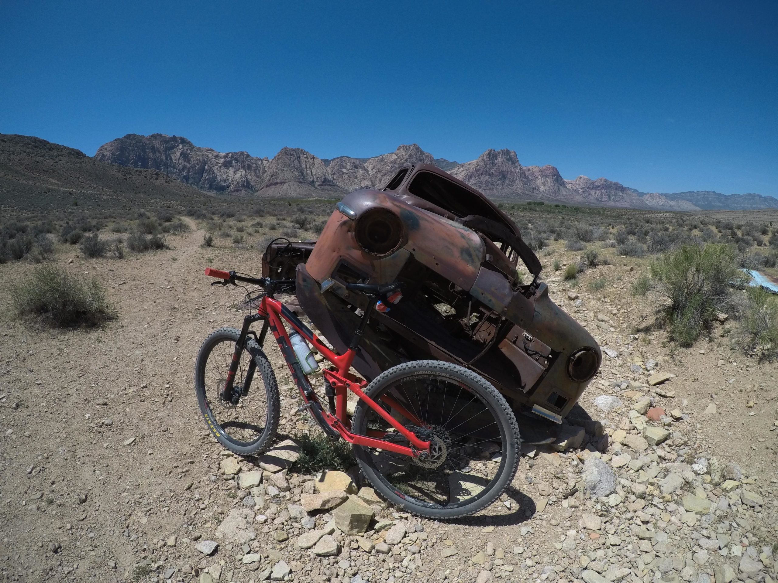 A red mountain bike parked next to a rusty, abandoned vehicle in a desert landscape. In the background, rugged mountains rise under a clear blue sky, with sparse vegetation surrounding the area. Blue Diamond mountain bike trail.