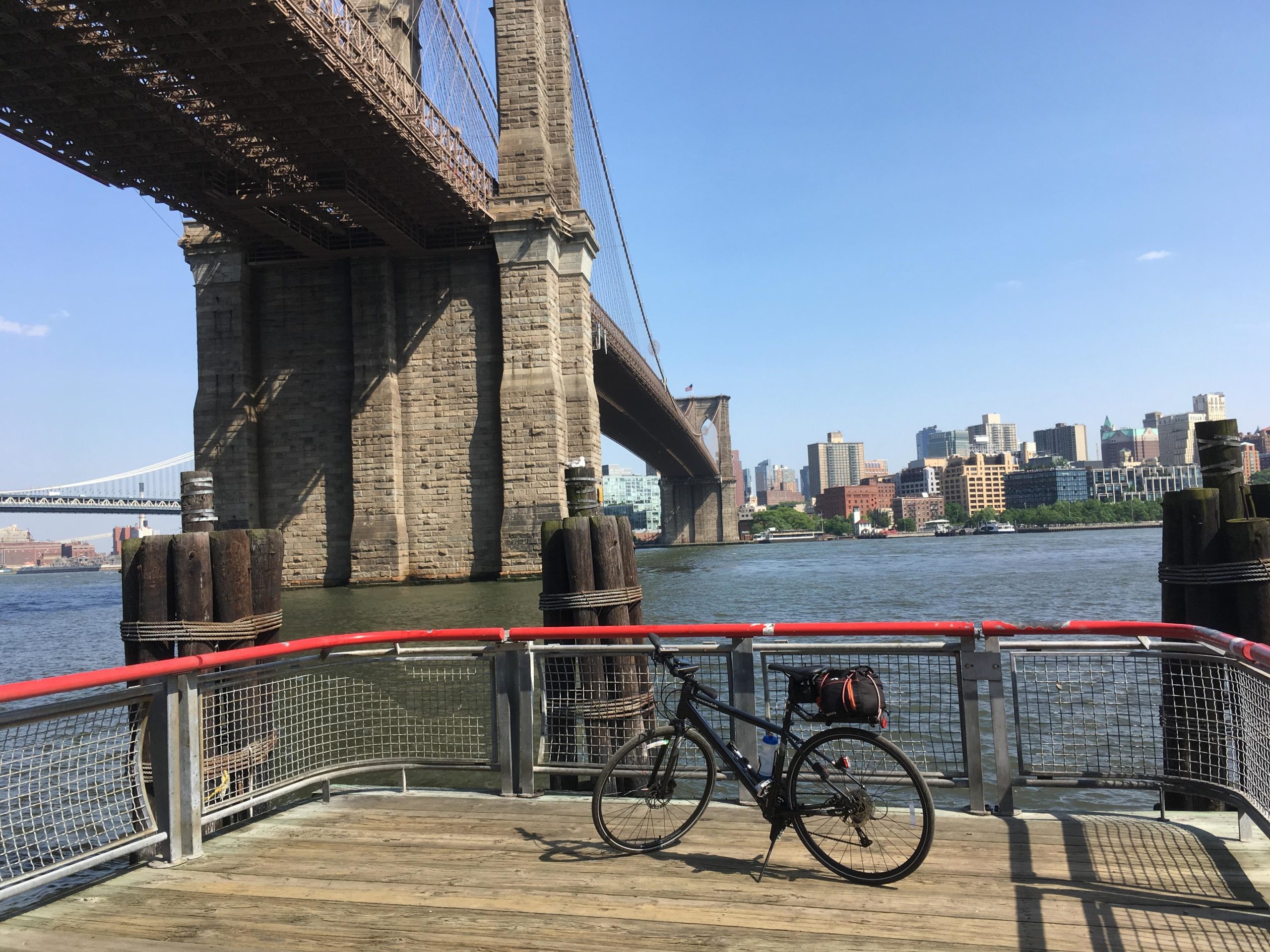 A bike is parked on a wooden pier with a red railing, overlooking a river. The view features the towering pillars of the Brooklyn Bridge and a skyline of city buildings in the background under a clear blue sky. East Side Green way 34th st to the Staten Island Ferry mountain bike trail.