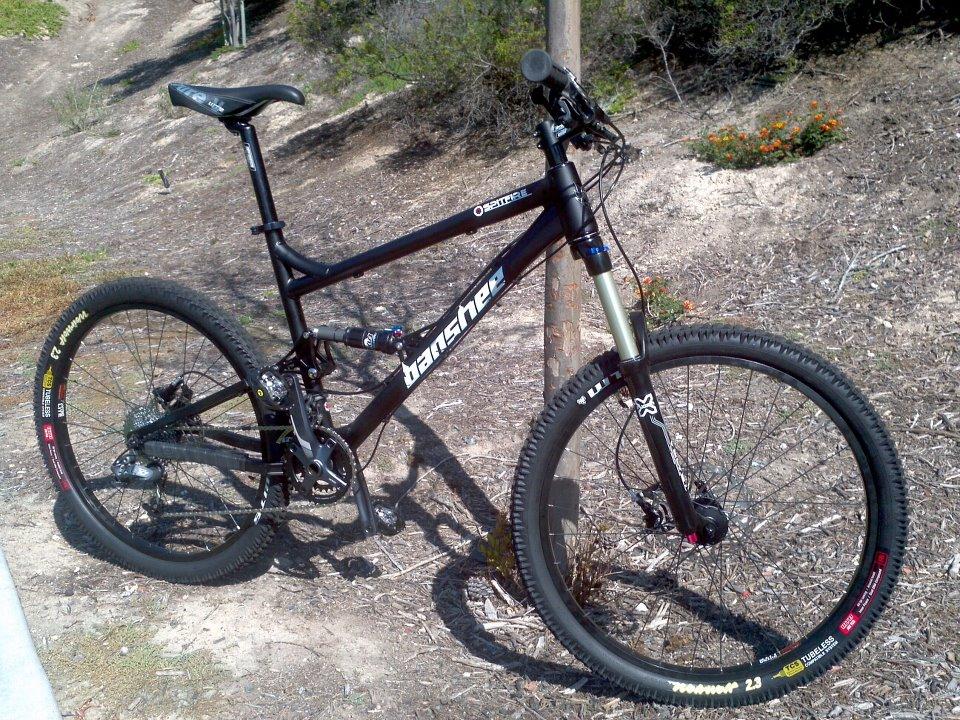 Banshee Spitfire: A black mountain bike leaning against a metal pole, showcasing its dual suspension system and knobby tires. The bike has a sleek design with the brand name "Banshee" visible on the frame. The background features a natural setting with dry grass and shrubs.