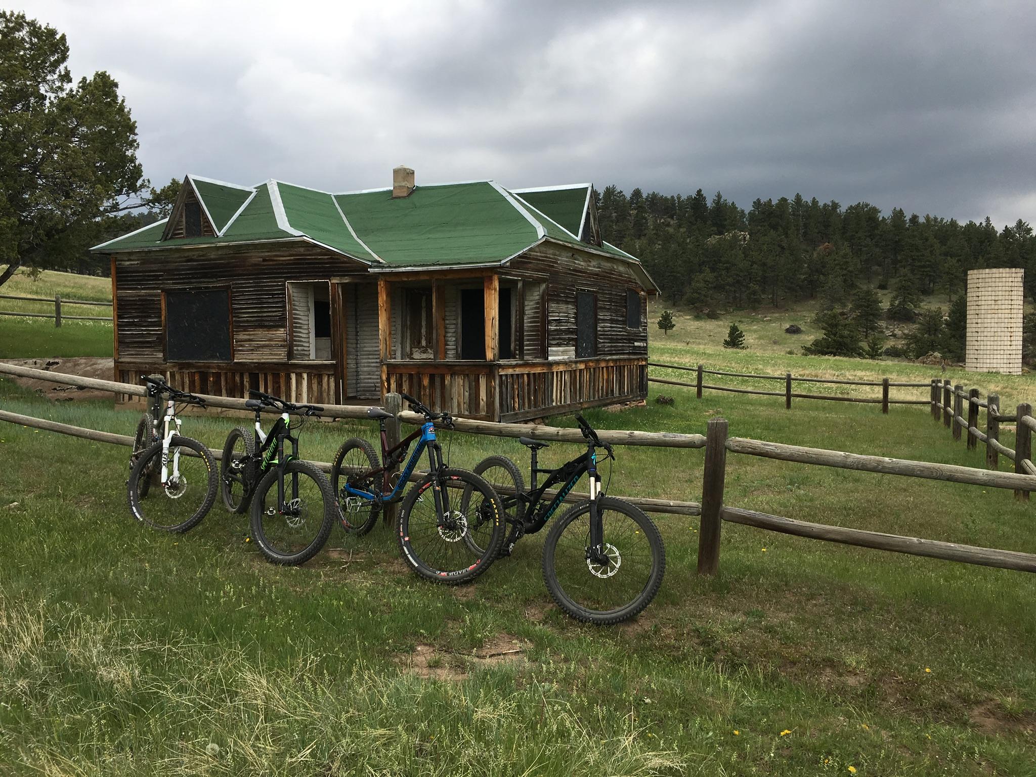 A rustic wooden house with a green roof is set in a grassy field, surrounded by trees and under a cloudy sky. In the foreground, four mountain bikes are parked along a wooden fence in front of the house. A silo can be seen in the distance. Hall Ranch mountain bike trail.