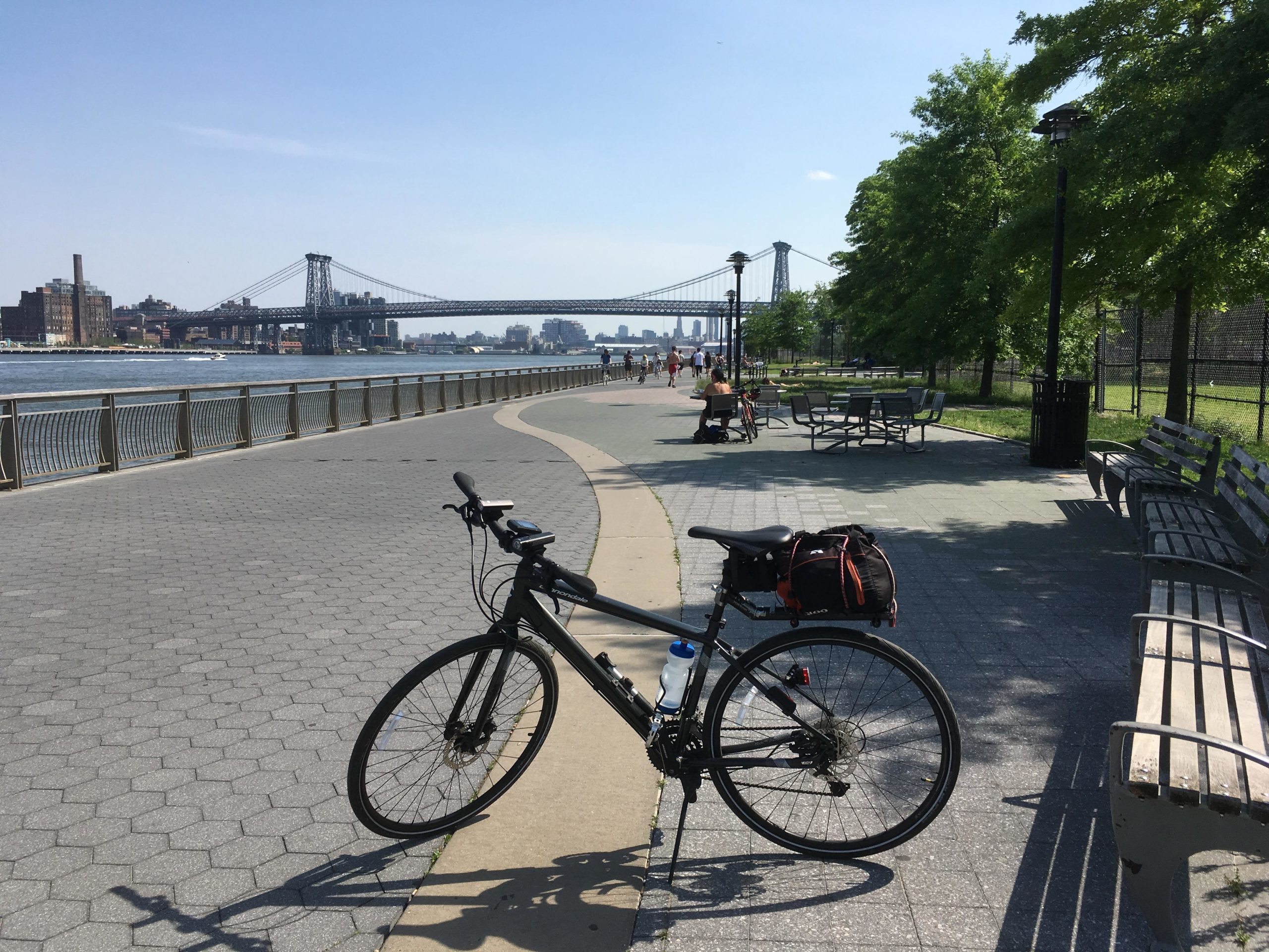 A black bicycle is parked on a paved path along a riverfront, with the Williamsburg Bridge visible in the background. The scene is sunny, featuring trees and benches lining the path. People can be seen walking and enjoying the waterfront area. East Side Green way 34th st to the Staten Island Ferry mountain bike trail.