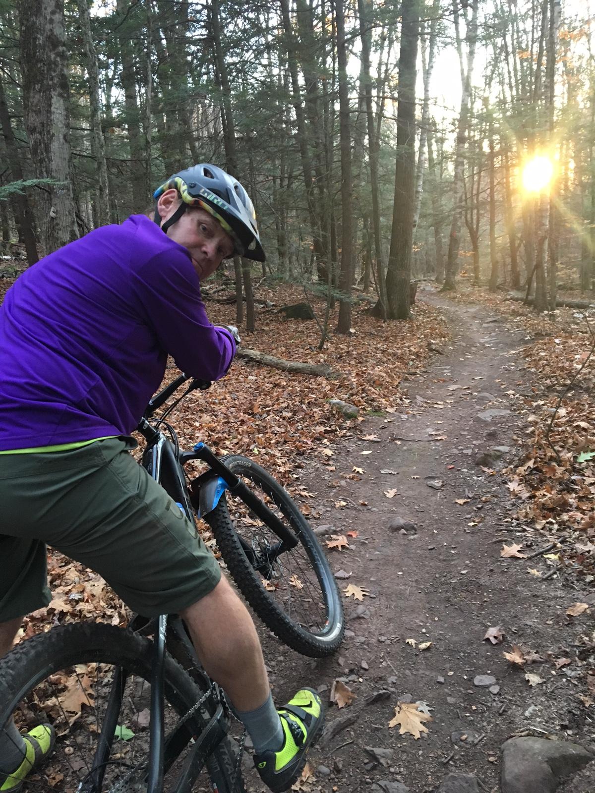 A person wearing a purple sweatshirt, green shorts, and a helmet is leaning on a mountain bike along a forest trail covered in autumn leaves. The sun is setting in the background, casting warm light through the trees, which are densely packed in the surroundings. Elm Ridge mountain bike trail.