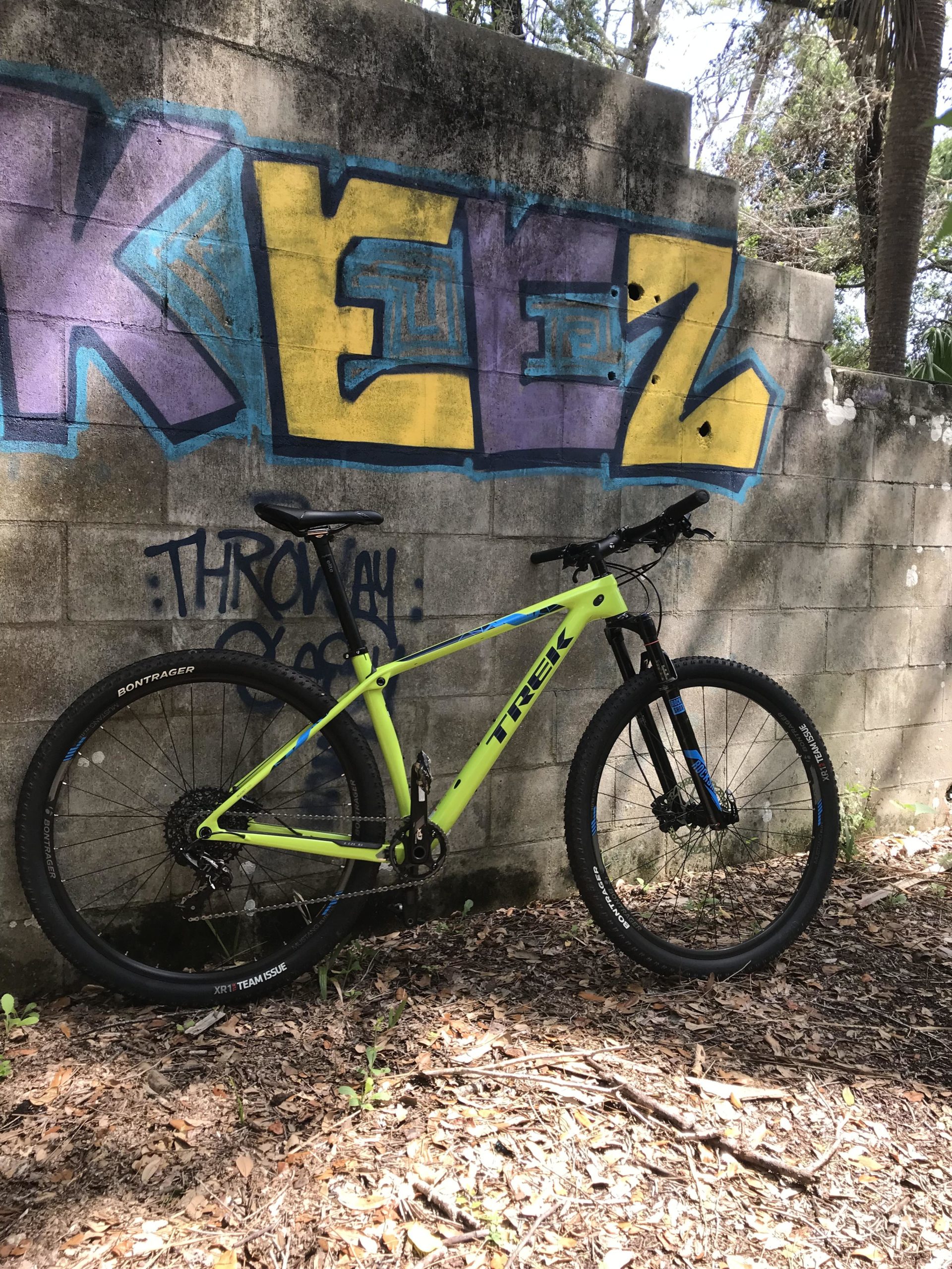 A bright lime green Trek mountain bike leaned against a graffiti-covered concrete wall. The graffiti features the word "KEEZ" in bold purple and yellow letters. Surrounding the bike and wall, there are patches of dirt and scattered leaves, with trees visible in the background. Mala Compra mountain bike trail.